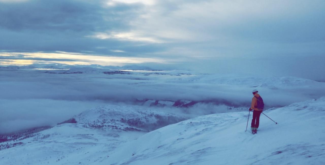 A skier in an orange jacket stands on a snowy slope, looking out over a vast, mountainous landscape covered in snow and fog under a cloudy sky.