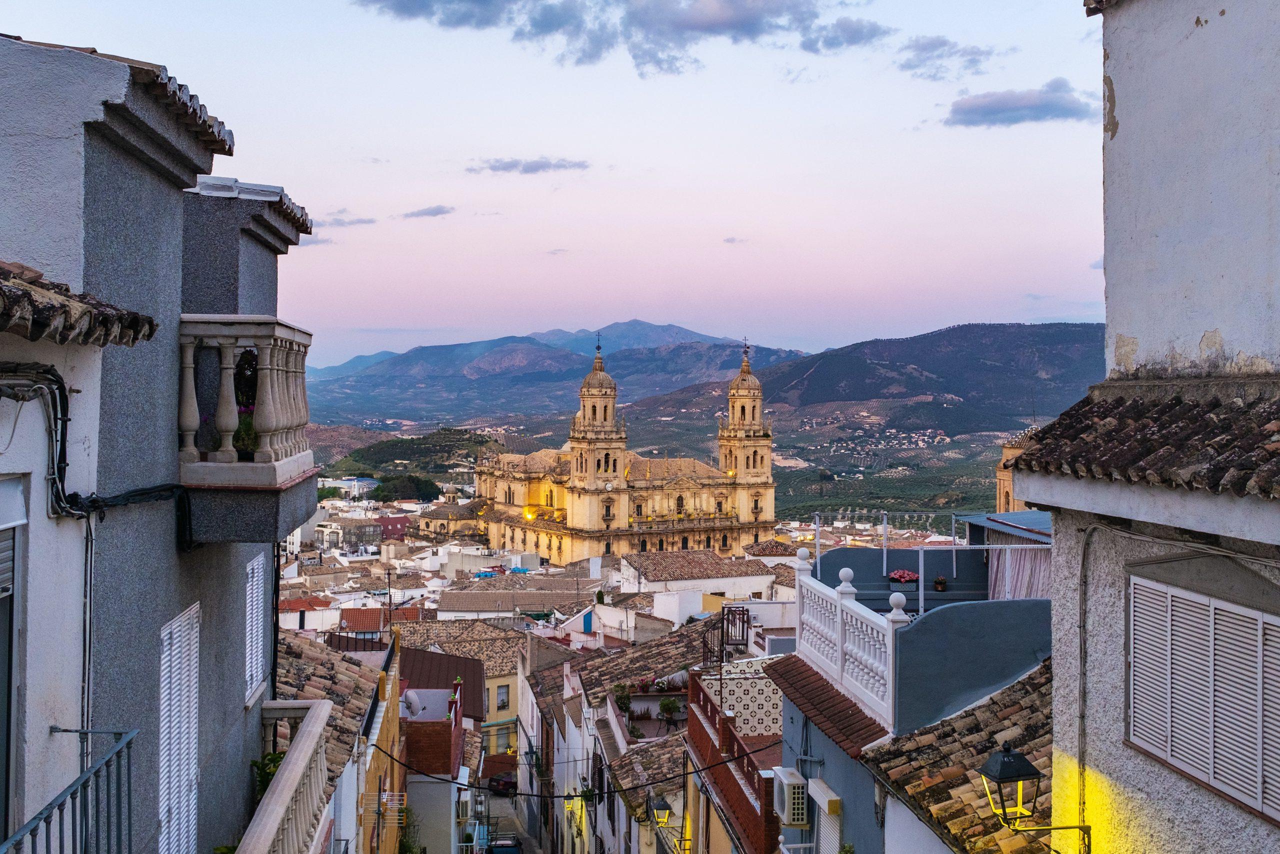 Ein Blick durch eine schmale Straße auf eine große, beleuchtete Kathedrale mit Zwillingstürmen in einer spanischen Stadt bei Sonnenuntergang, umgeben von weißen Gebäuden mit Ziegeldächern und fernen Bergen unter einem sanften, pastellfarbenen Himmel.