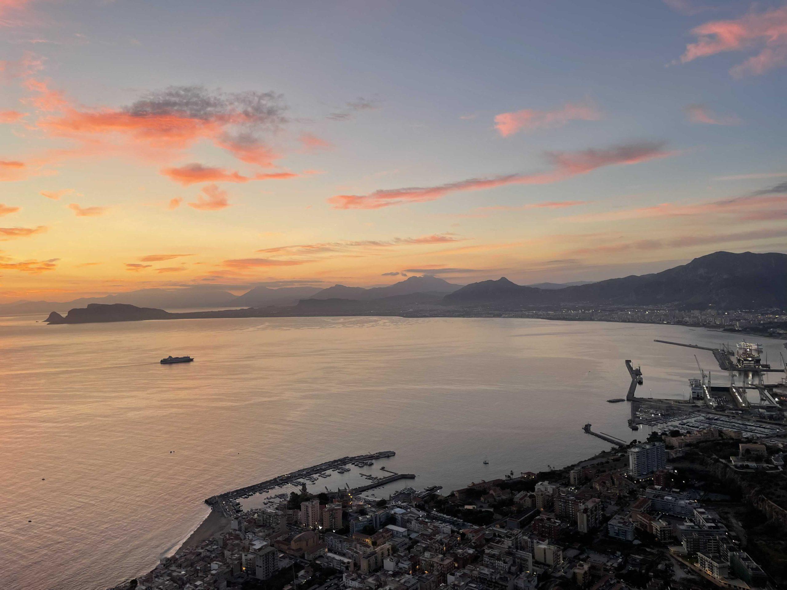 Eine Küstenstadt bei Sonnenuntergang mit bunten Wolken, Bergen im Hintergrund, einem Schiff in der Bucht und einem Hafen mit Docks und Gebäuden entlang der Küstenlinie.