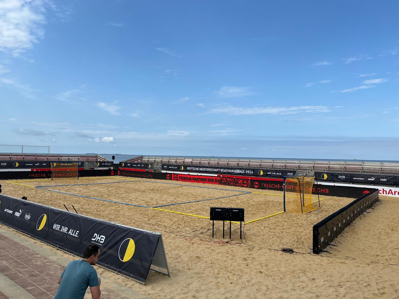 Ein sandiges, leeres Beachhandballfeld mit schwarzen Barrieren und gelben Torpfosten ist am Meer unter blauem Himmel aufgebaut. Eine Person steht im Vordergrund neben dem Spielfeld.
