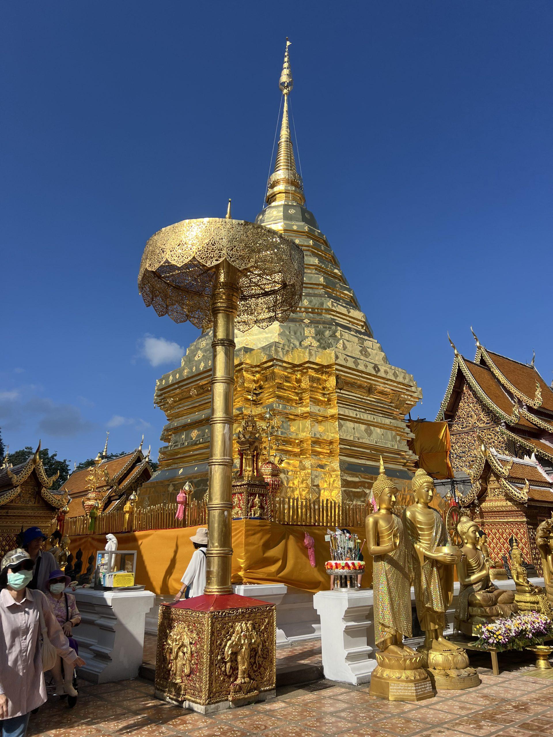 Ein goldener buddhistischer Stupa, umgeben von Statuen und kunstvollen Verzierungen, unter einem klaren blauen Himmel, mit Menschen, die auf dem Tempelgelände spazieren gehen und beten.