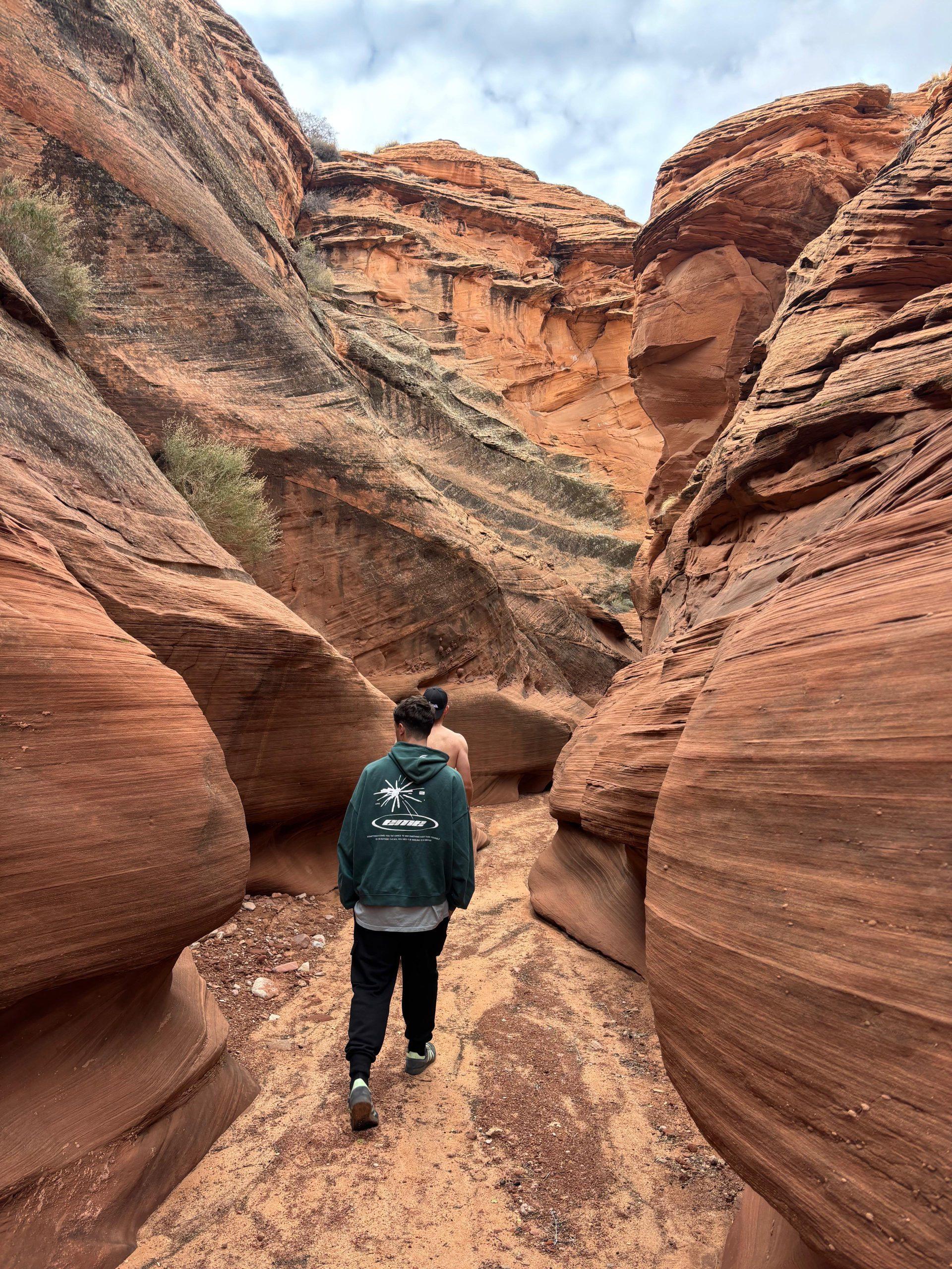 Zwei Personen gehen unter bewölktem Himmel durch einen engen, gewundenen Slot Canyon mit hoch aufragenden roten und orangefarbenen Felswänden. Eine Person trägt eine grüne Jacke und die andere eine Duttfrisur.