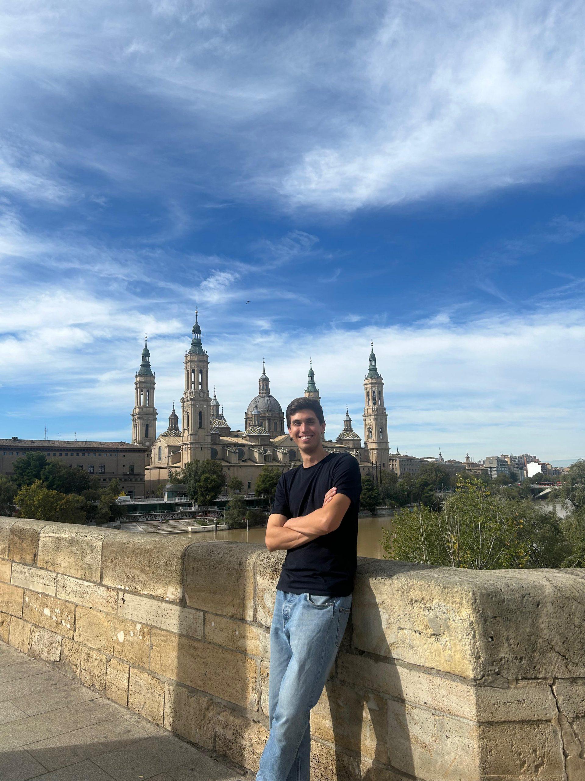 Eine Person in schwarzem Hemd und blauer Jeans steht lächelnd und mit verschränkten Armen auf einer Steinbrücke, im Hintergrund eine große Kathedrale mit mehreren Türmen und Kuppeln unter einem strahlend blauen Himmel.