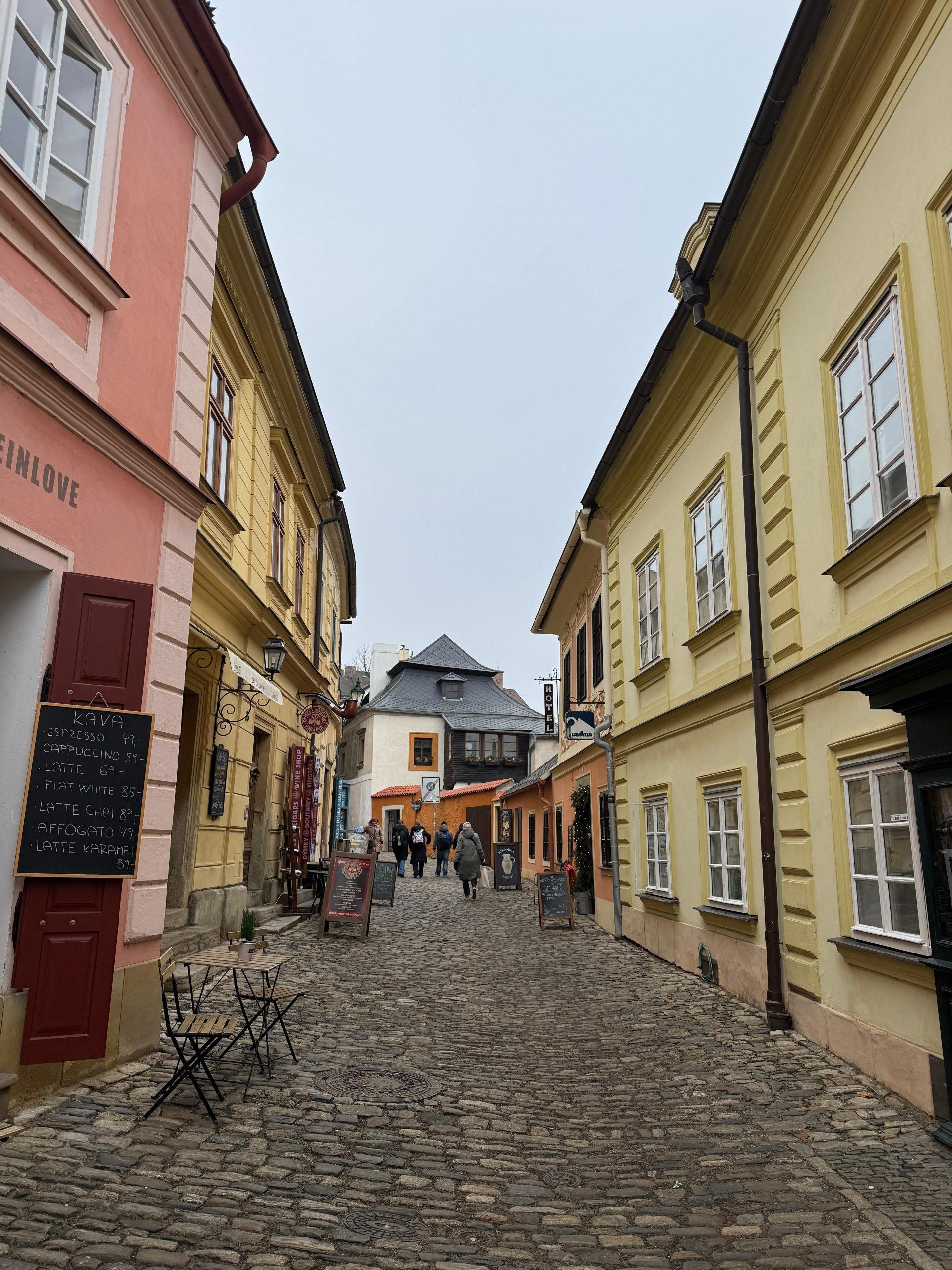 Eine schmale Kopfsteinpflasterstraße mit pastellgelben und rosafarbenen Gebäuden, auf der linken Seite Tische eines Straßencafés und in der Ferne Gruppen von Spaziergängern unter einem bedeckten Himmel.