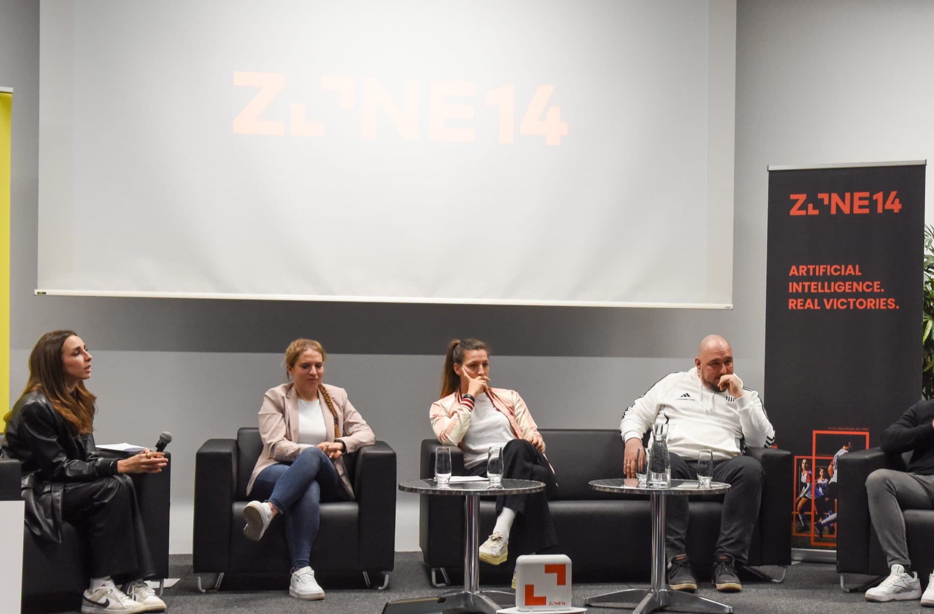 Fünf Personen sitzen auf einer Bühne und nehmen an einer Podiumsdiskussion teil. Ein Mann steht auf und spricht in ein Mikrofon. Hinter ihnen sind Transparente mit den Aufschriften "ZONE14" und "Künstliche Intelligenz. Echte Siege.".