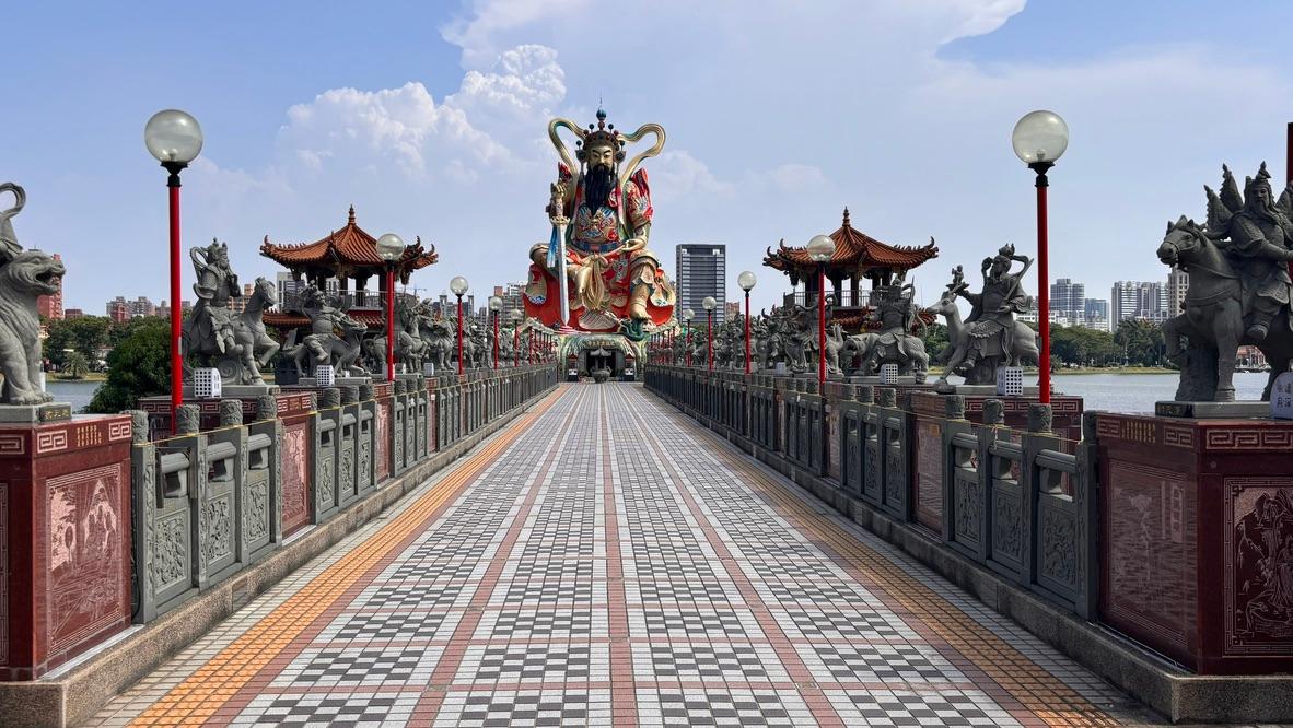 Eine lange, geflieste Brücke mit Statuen und Laternen führt zu einer großen, farbenfrohen Götterstatue am Ende, mit Stadtgebäuden und einem teilweise bewölkten Himmel im Hintergrund.