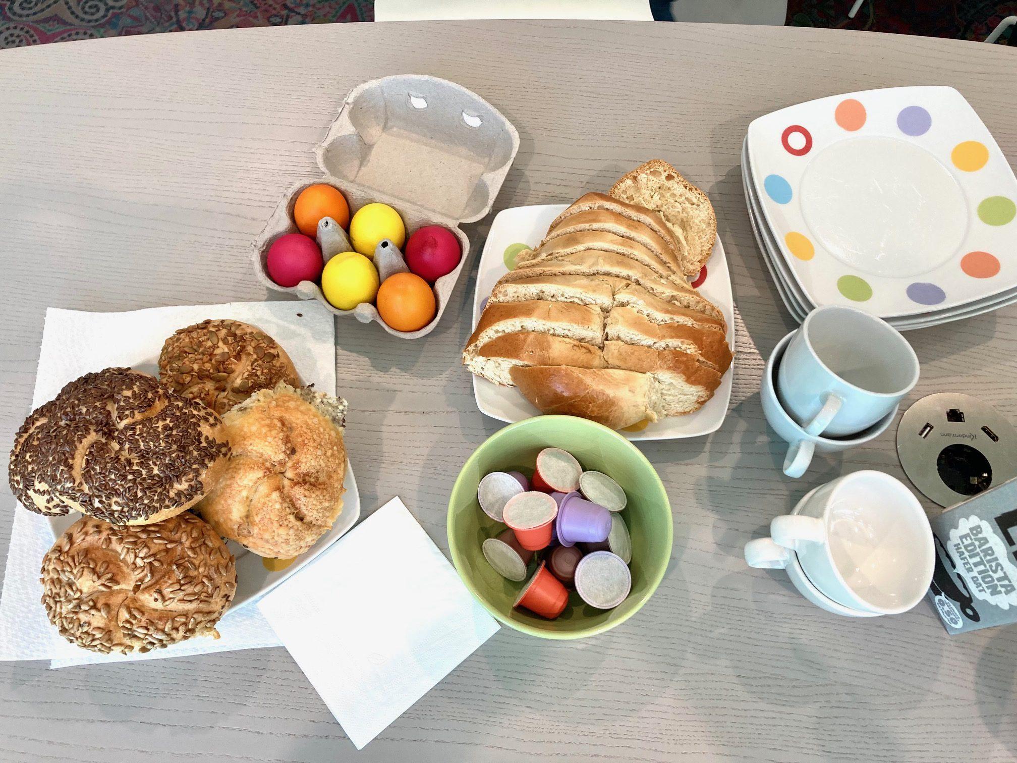 Ein Frühstücksbuffet mit verschiedenen Brötchen, geschnittenem Brot, bunten Eiern in einem Karton, einer Schale mit Kaffeesahne, gestapelten Tellern und Tassen auf einem hellen Holztisch.
