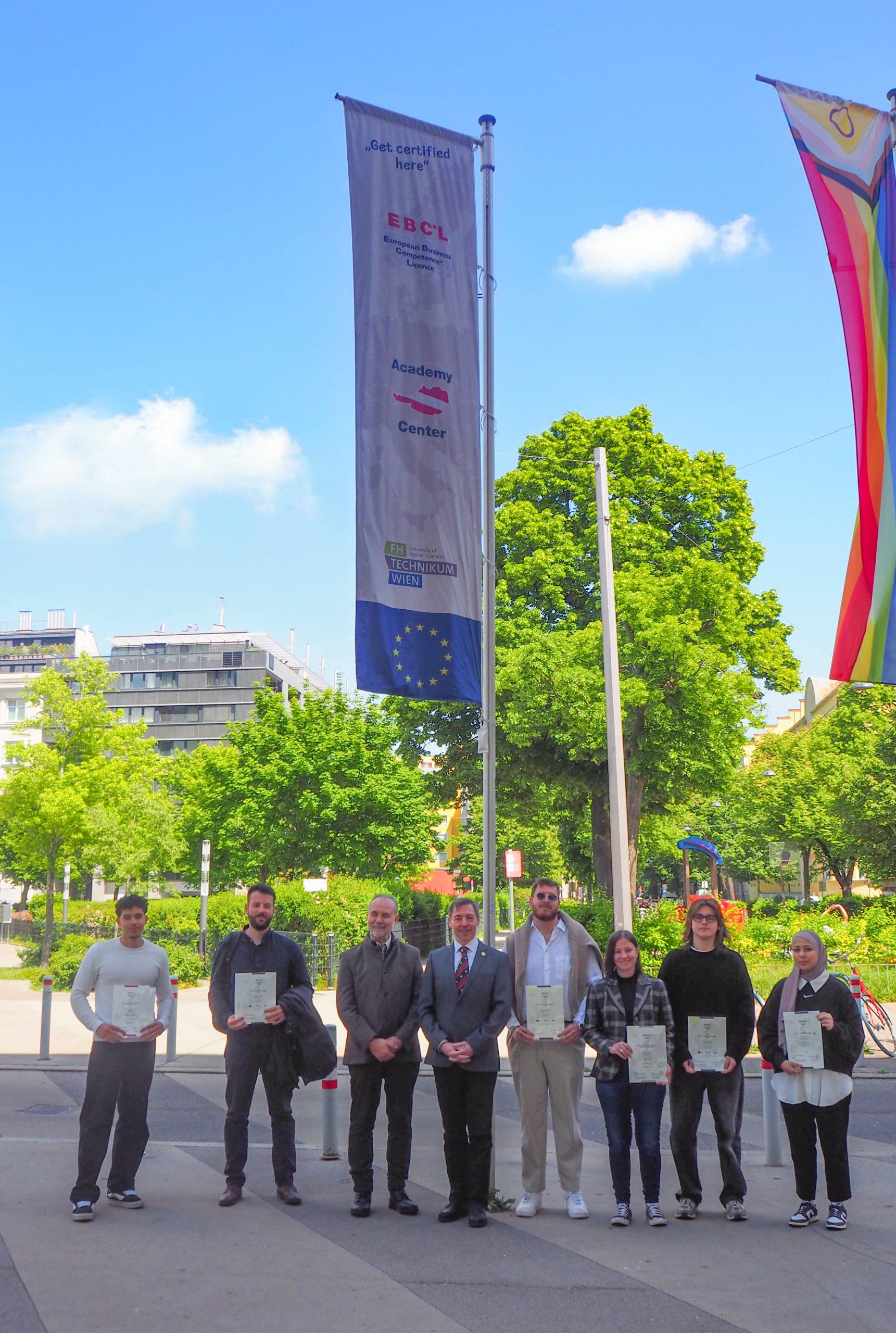 Acht Personen stehen in einer Reihe im Freien und halten Urkunden in der Hand. Hinter ihnen stehen hohe Fahnenmasten mit Bannern und Flaggen, grüne Bäume und moderne Gebäude unter einem strahlend blauen Himmel.