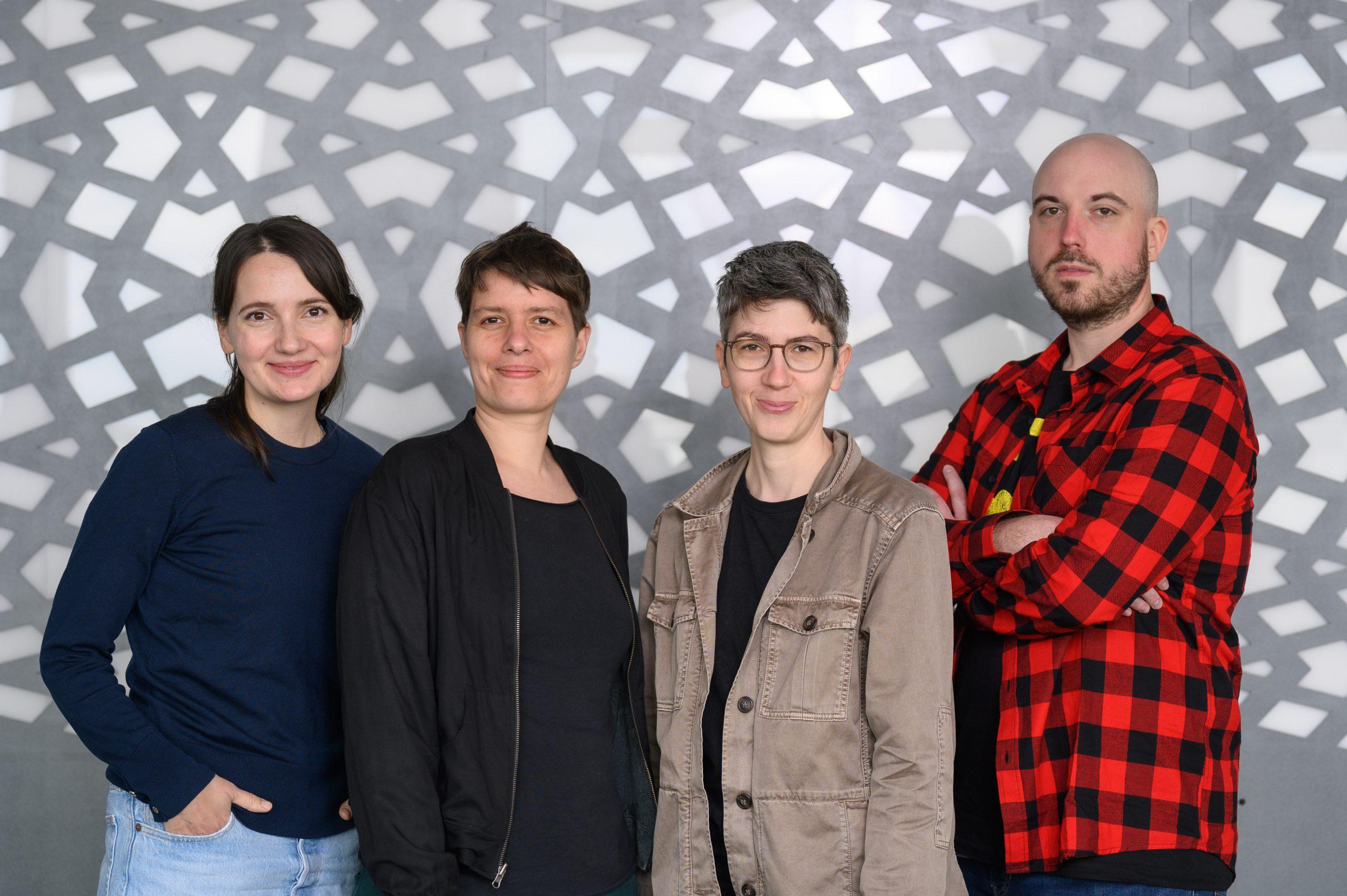 Vier Erwachsene stehen nebeneinander, lächeln in die Kamera und stehen vor einer grau-weiß gemusterten Wand. Drei von ihnen sind Frauen in legeren Jacken, einer ist ein Mann in einem rotkarierten Hemd mit verschränkten Armen.