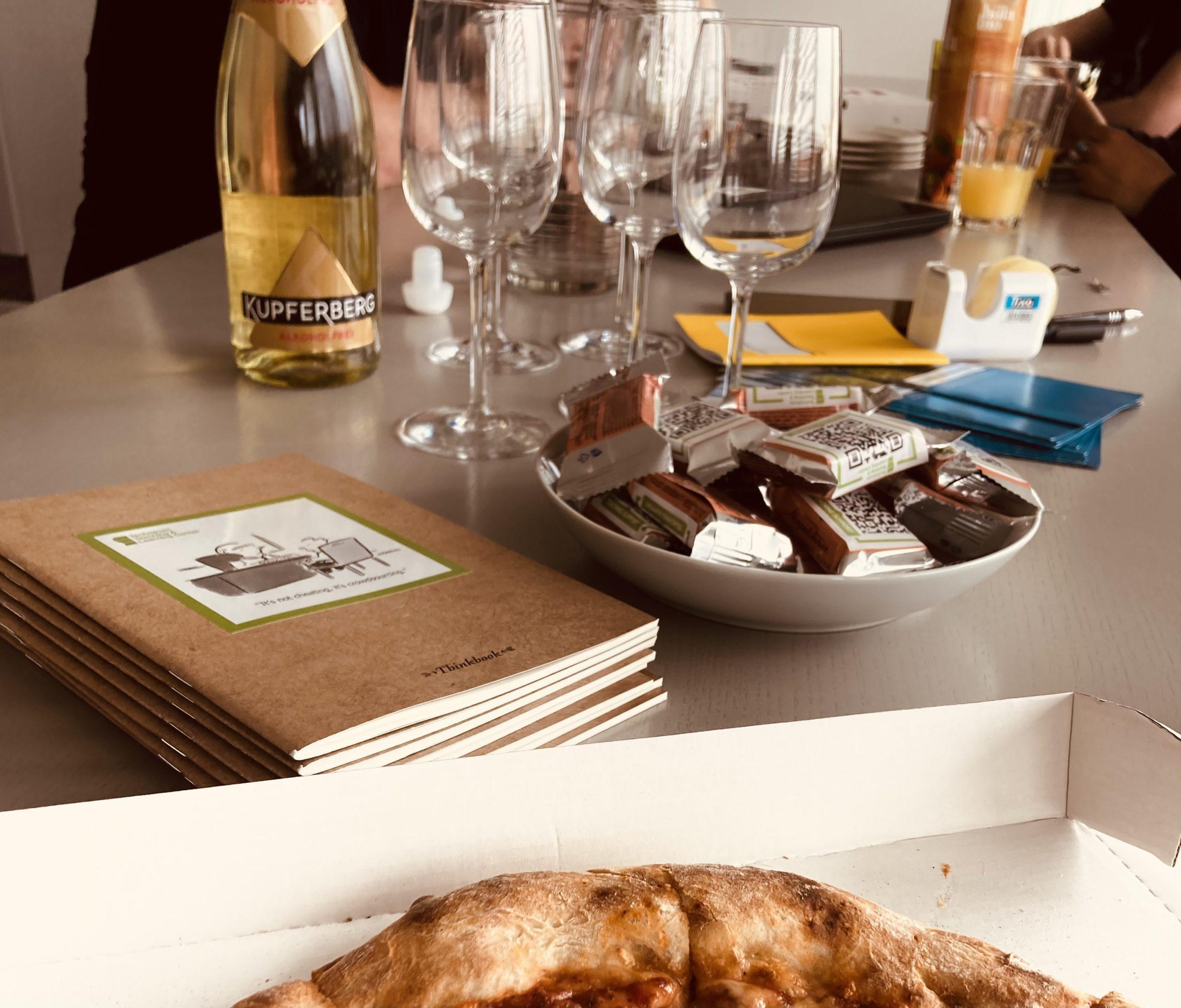 Ein Tisch mit einem Stapel Notizbücher, einer Schale mit kleinen Schokoriegeln, einer Pizza in einer Schachtel, einer Flasche Wein, mehreren leeren Weingläsern und Büromaterial. Im Hintergrund sind teilweise Menschen zu sehen.