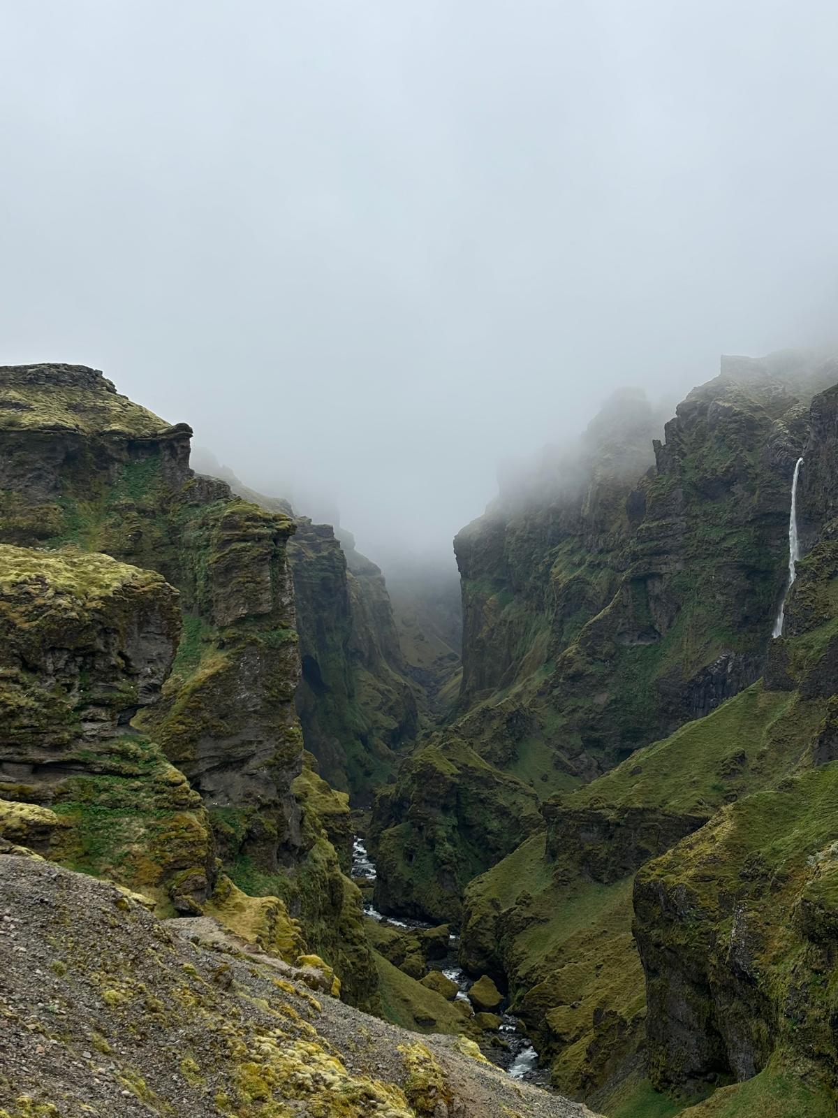 Eine tiefe, grüne Schlucht mit steilen, moosbewachsenen Felsen und einem kleinen Fluss, der sie durchfließt, teilweise in dichten Nebel gehüllt und unter einem bewölkten Himmel.