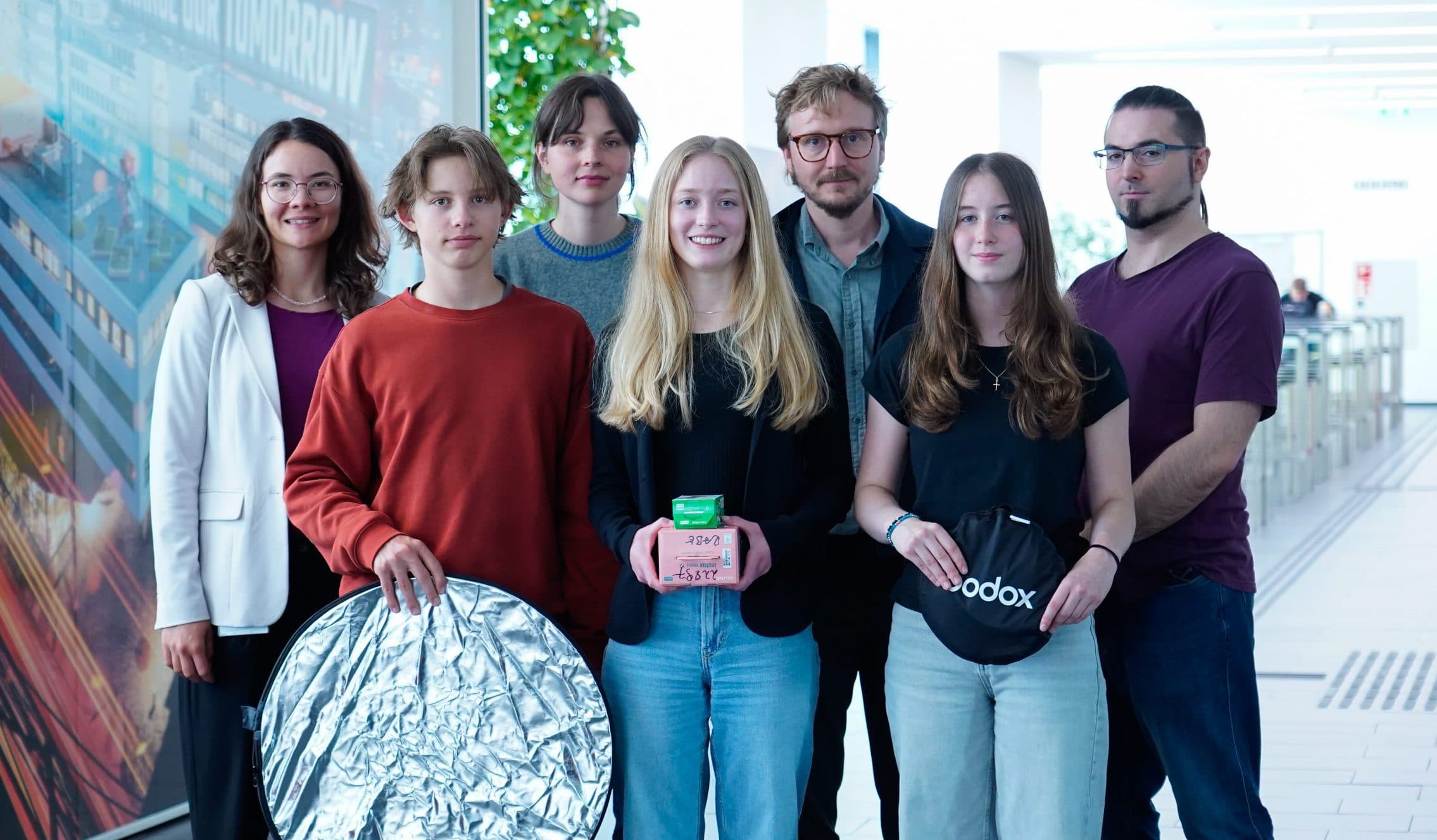 Eine Gruppe von sieben Personen, vier Frauen und drei Männer, steht in einem Raum und lächelt. Zwei halten eine Fotoausrüstung, darunter einen silbernen Reflektor und eine Godox-Softbox. Der Hintergrund zeigt einen hellen Flur und ein großes Wandgemälde.