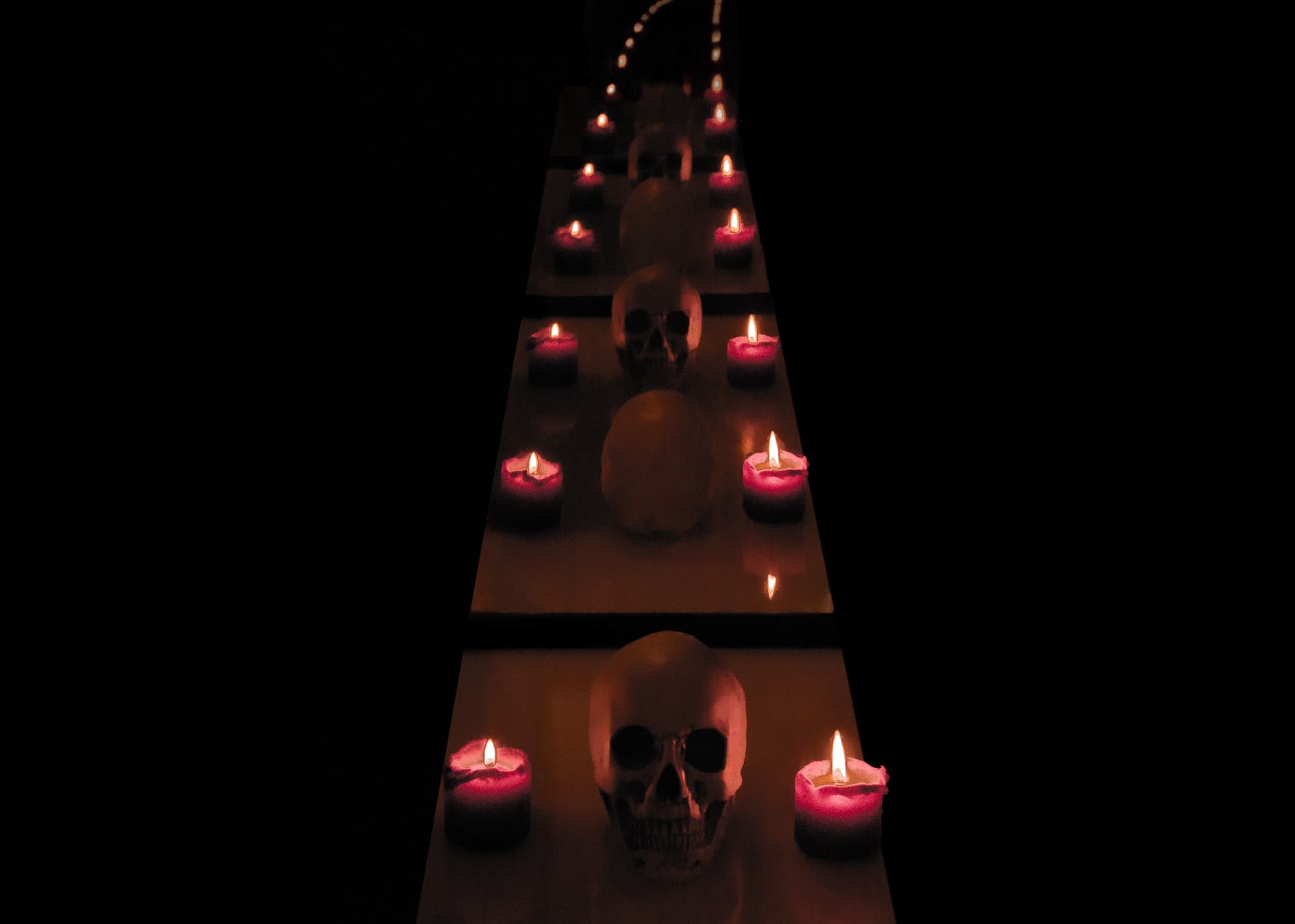 Eine Reihe menschlicher Schädelnachbildungen und brennende rote Kerzen sind abwechselnd auf einer spiegelnden Oberfläche angeordnet und erzeugen einen dramatischen, symmetrischen Effekt in einer dunklen Umgebung.