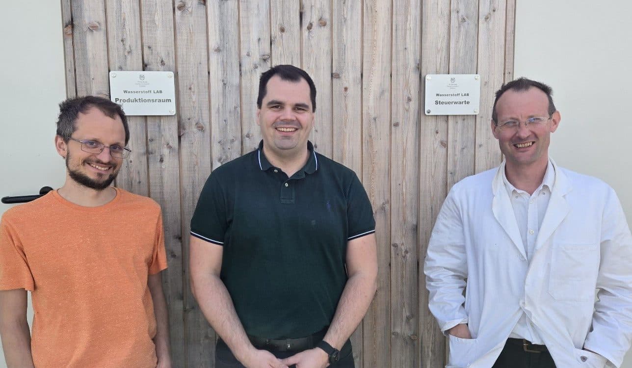 Drei Männer stehen lächelnd vor einer Holzwand mit zwei Plaketten. Der Mann auf der linken Seite trägt eine Brille und ein orangefarbenes Hemd, der Mann in der Mitte ein schwarzes Polohemd und der Mann auf der rechten Seite eine Brille und einen weißen Mantel.