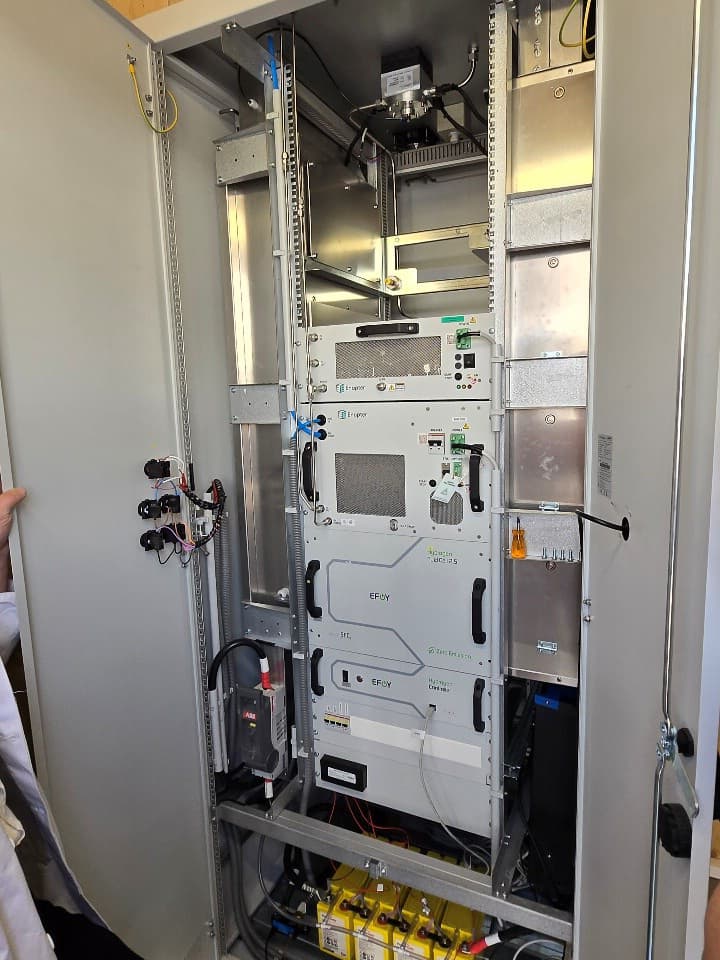 In einem Metallschrank, dessen Türen geöffnet sind, stapeln sich verschiedene elektronische Geräte, Kabel und Überwachungsvorrichtungen, darunter Bildschirme, Knöpfe und Stecker, die wahrscheinlich für technische oder wissenschaftliche Zwecke verwendet werden.