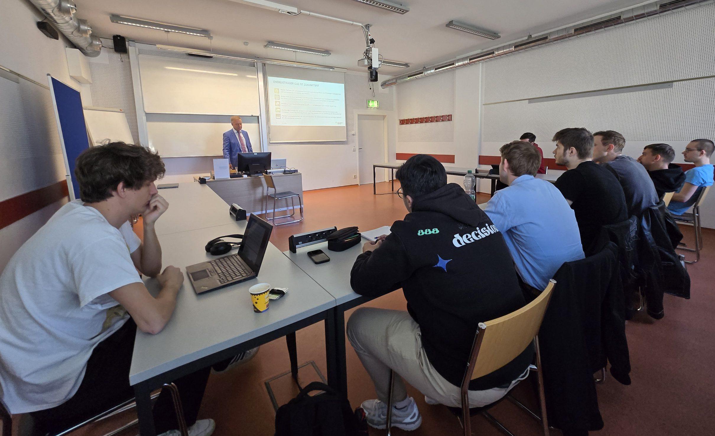 Eine Gruppe von Studierenden sitzt an Tischen und blickt auf einen Dozenten, der neben einem Bildschirm steht, auf dem eine Präsentation in einem Klassenzimmer gezeigt wird. Laptops und Notebooks liegen auf den Tischen.