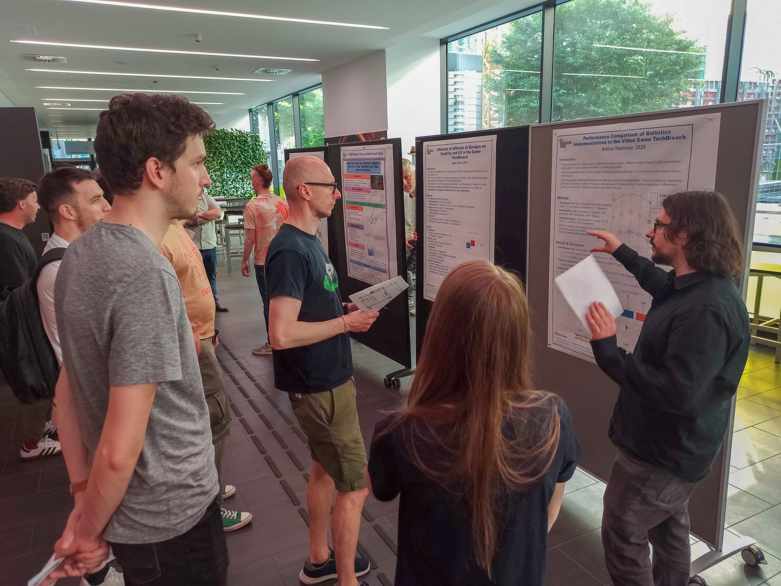 Eine Gruppe von Menschen steht in einem Innenraum um einen Vortragenden herum, der ein wissenschaftliches Poster auf einer Tafel erklärt, während andere zuhören und Dokumente halten. Große Fenster zeigen Bäume und städtische Gebäude draußen.