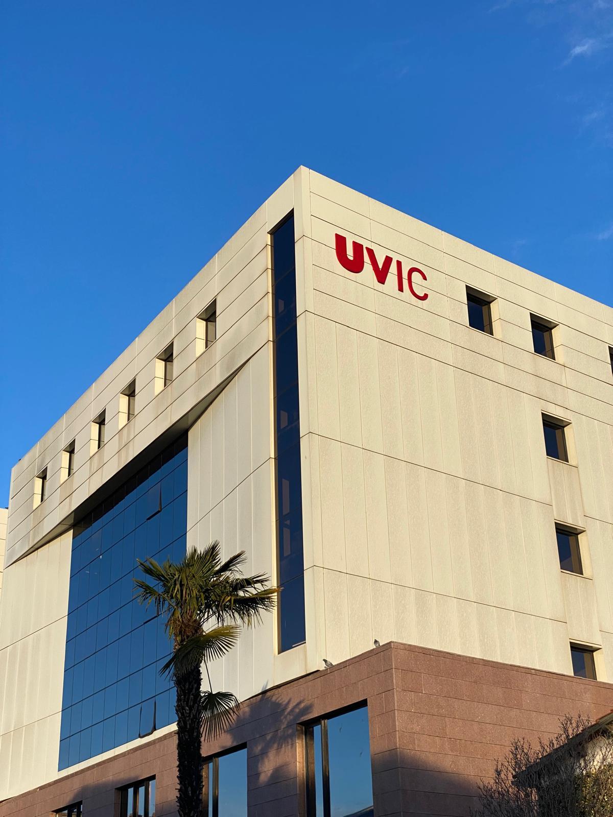 Ein modernes beigefarbenes Gebäude mit großen Fenstern und den roten Buchstaben UVIC an der oberen Ecke, vor einem klaren blauen Himmel und einer Palme.