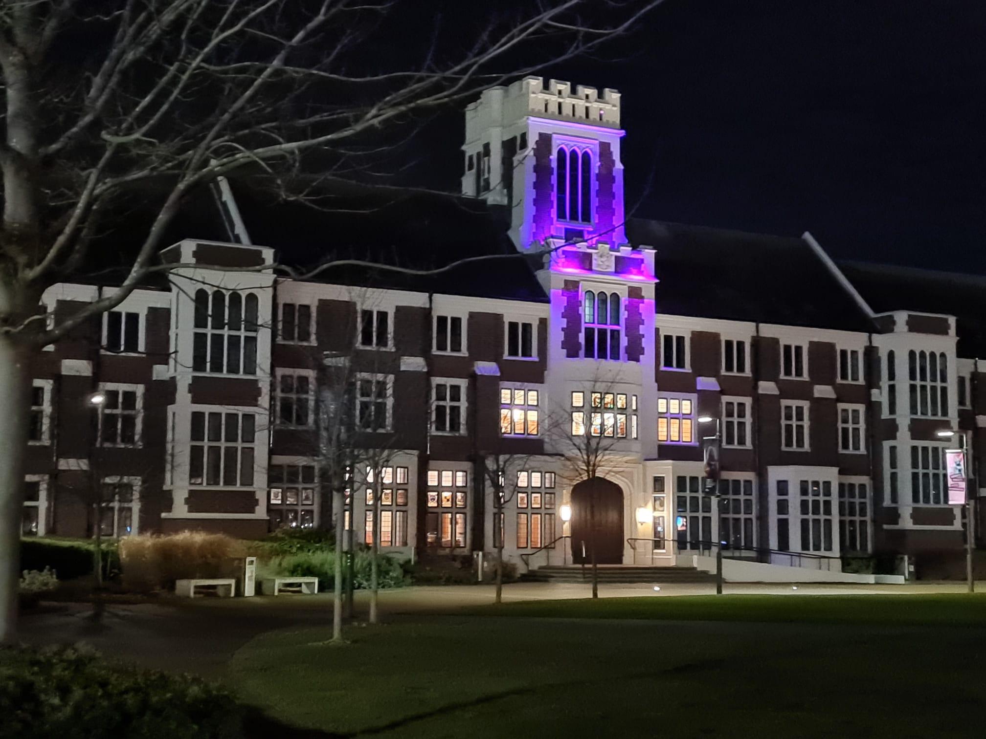 Ein großes, historisches Gebäude mit hohen Fenstern und einem zentralen Turm wird nachts von violetten und gelben Lichtern beleuchtet. Das Gebäude ist von Gras, Bäumen ohne Blätter und Wegen umgeben.