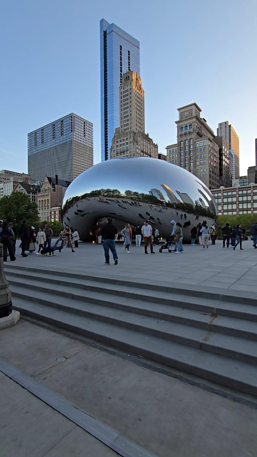 Menschen versammeln sich um Cloud Gate (The Bean), eine große reflektierende Skulptur im Millennium Park in Chicago, mit Wolkenkratzern und einem klaren Himmel im Hintergrund. Einige Besucher steigen die Stufen im Vordergrund hinauf.