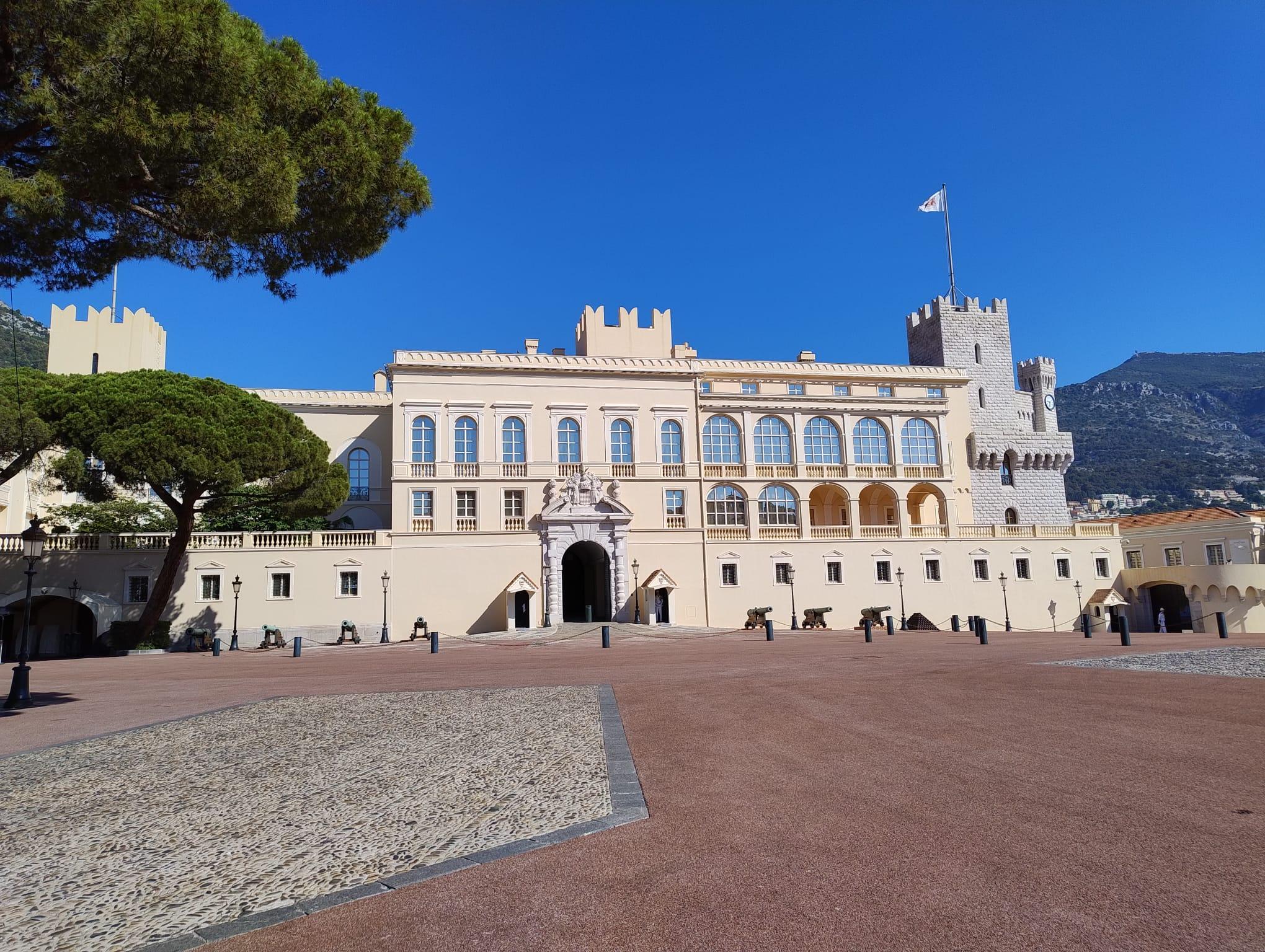 Das Bild zeigt das Äußere des Fürstenpalastes von Monaco, ein großes cremefarbenes Gebäude mit Bogenfenstern, Zinnen und einem zentralen Turm vor einem klaren blauen Himmel und einem bergigen Hintergrund.