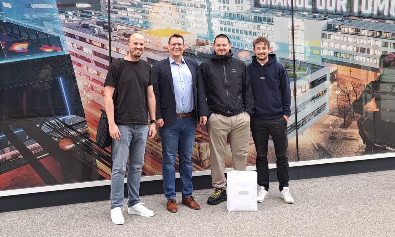 Vier Männer stehen lächelnd vor einem großen Wandbild an der FH Technikum Wien, das urbane und technologische Bilder und den Slogan CHANGE OUR TOMORROW zeigt.