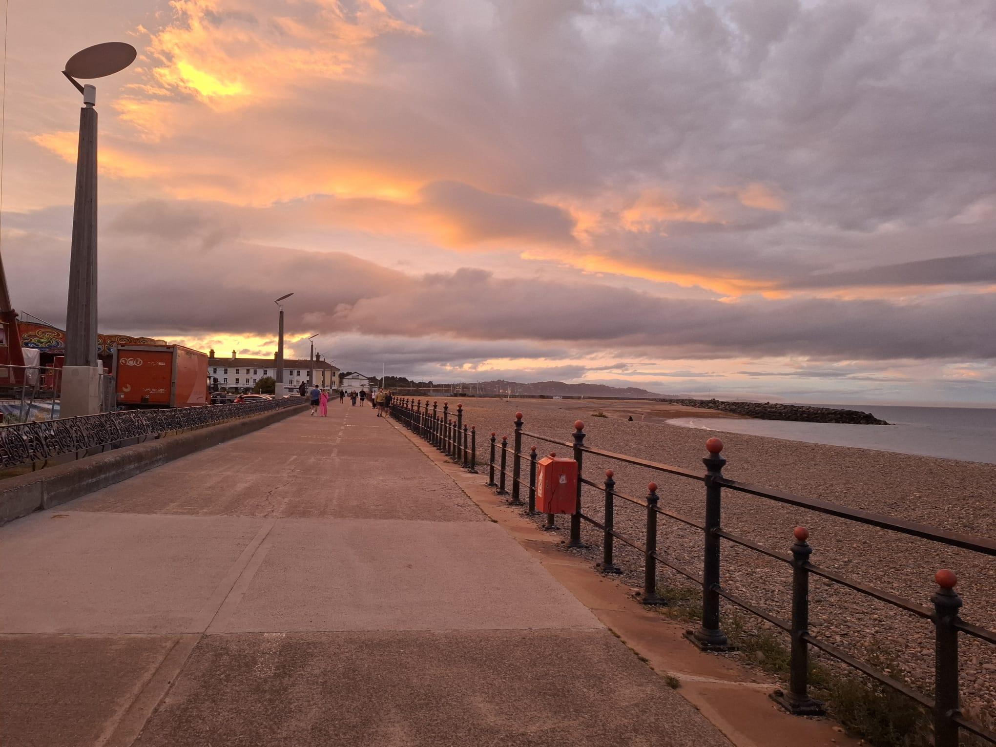 Eine breite Promenade entlang eines Kieselstrandes bei Sonnenuntergang, mit dramatischen orangefarbenen Wolken am Himmel. Laternenpfähle säumen den Weg und ein paar Menschen gehen in der Ferne in der Nähe von weißen Gebäuden am Meer spazieren.