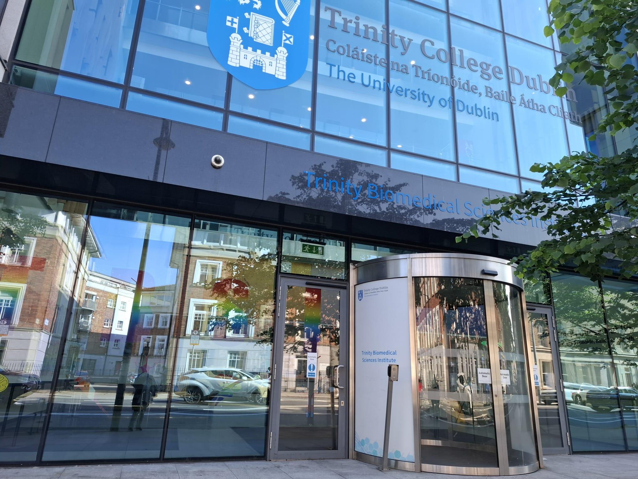 Gläserner Eingang des Trinity Biomedical Sciences Institute am Trinity College Dublin, in dem sich die umliegenden Gebäude spiegeln, wobei der Name und das Logo der Universität auf der oberen Fassade zu sehen sind.