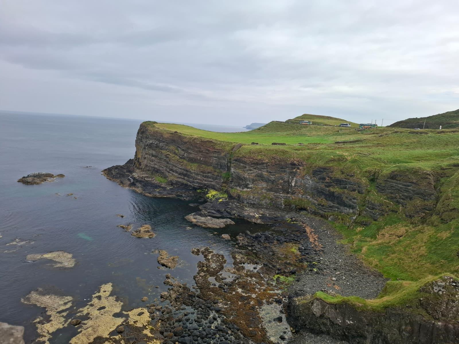 Eine felsige Küste mit steilen Klippen, die mit grünem Gras bewachsen sind, blickt auf ein ruhiges Meer unter einem bewölkten Himmel. Auf dem grasbewachsenen Plateau oberhalb der Klippen sind kleine Gebäude zu sehen.