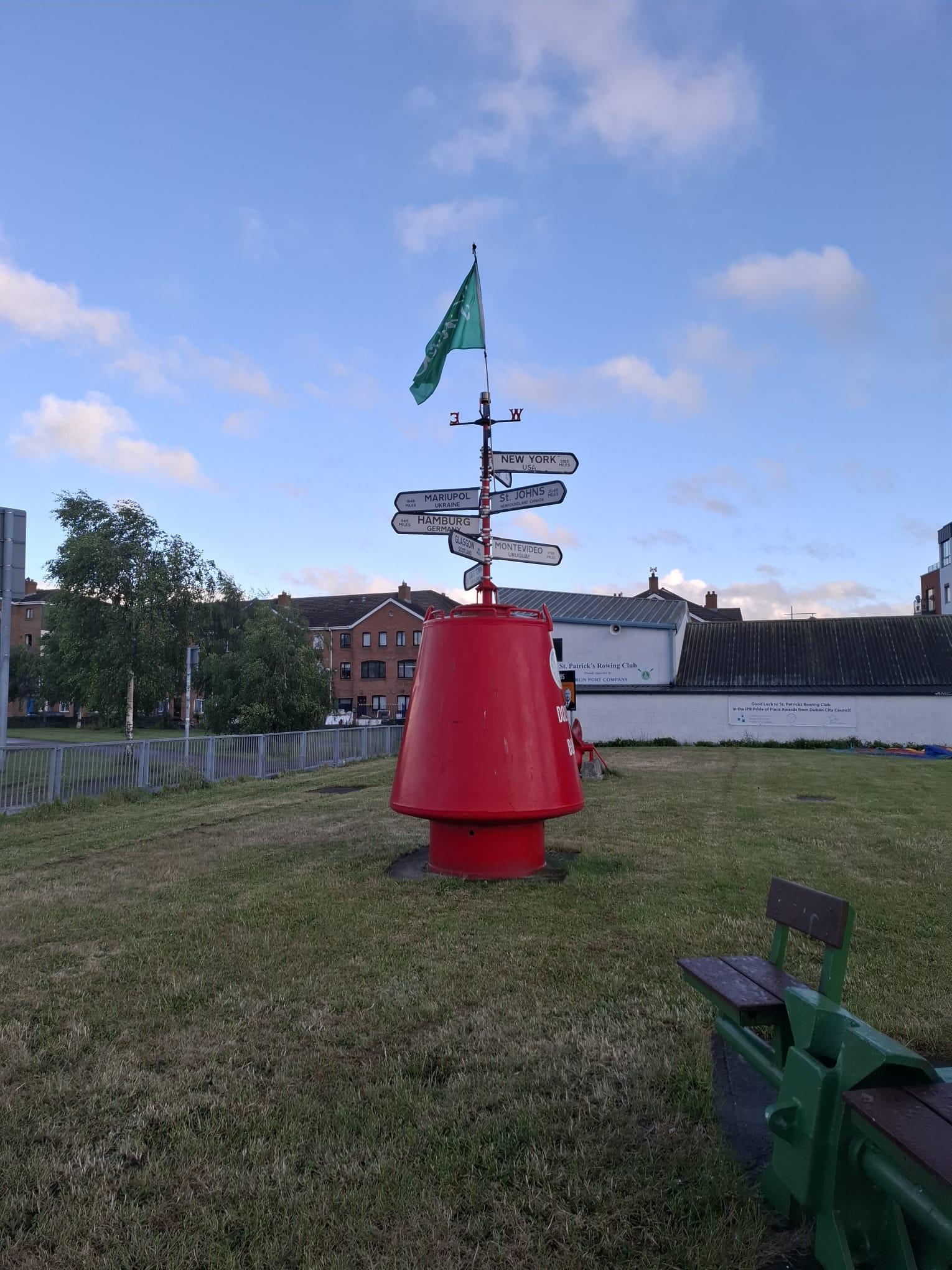 Eine große rote Boje mit einer grünen Flagge an der Spitze steht auf einer Grasfläche. Richtungsschilder weisen auf verschiedene Orte hin. Im Hintergrund sind Bänke und Gebäude unter einem teilweise bewölkten blauen Himmel zu sehen.