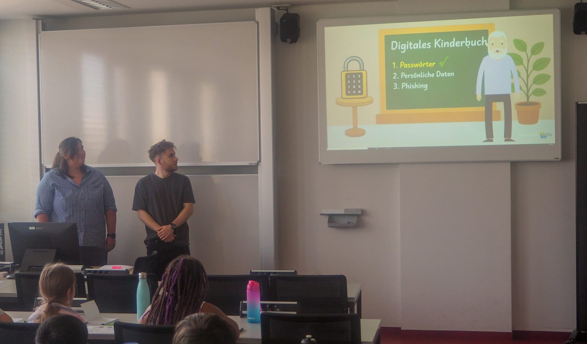 Zwei Erwachsene stehen vor einem Klassenzimmer und halten eine Präsentation für Kinder. Eine Folie auf dem Bildschirm mit dem Titel