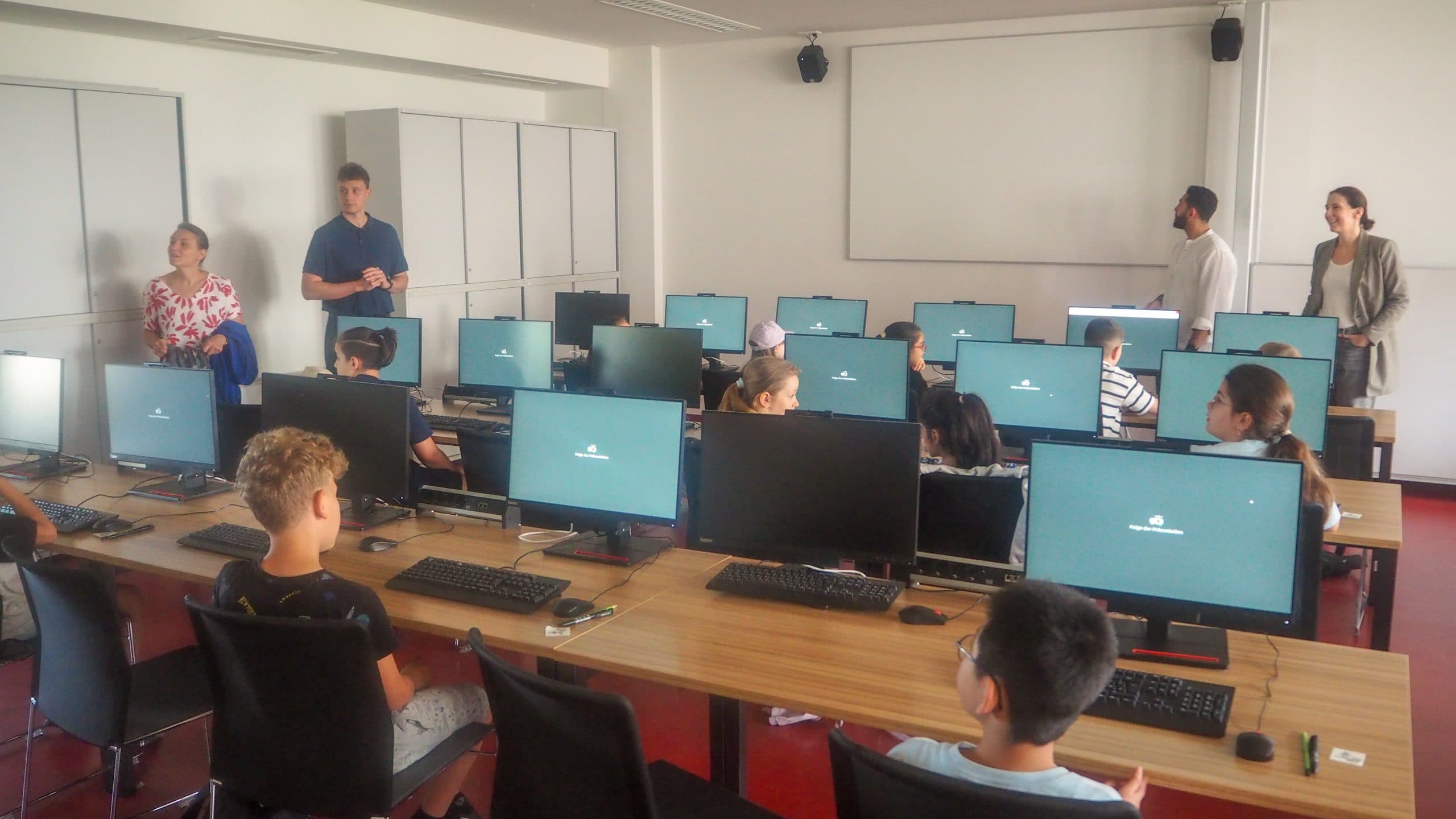 Die Kinder sitzen an Tischen mit Computermonitoren in einem Klassenzimmer, während die Erwachsenen im hinteren und seitlichen Teil des Raums stehen. Die Monitore zeigen einen Anmeldebildschirm an und der Raum ist hell und modern.