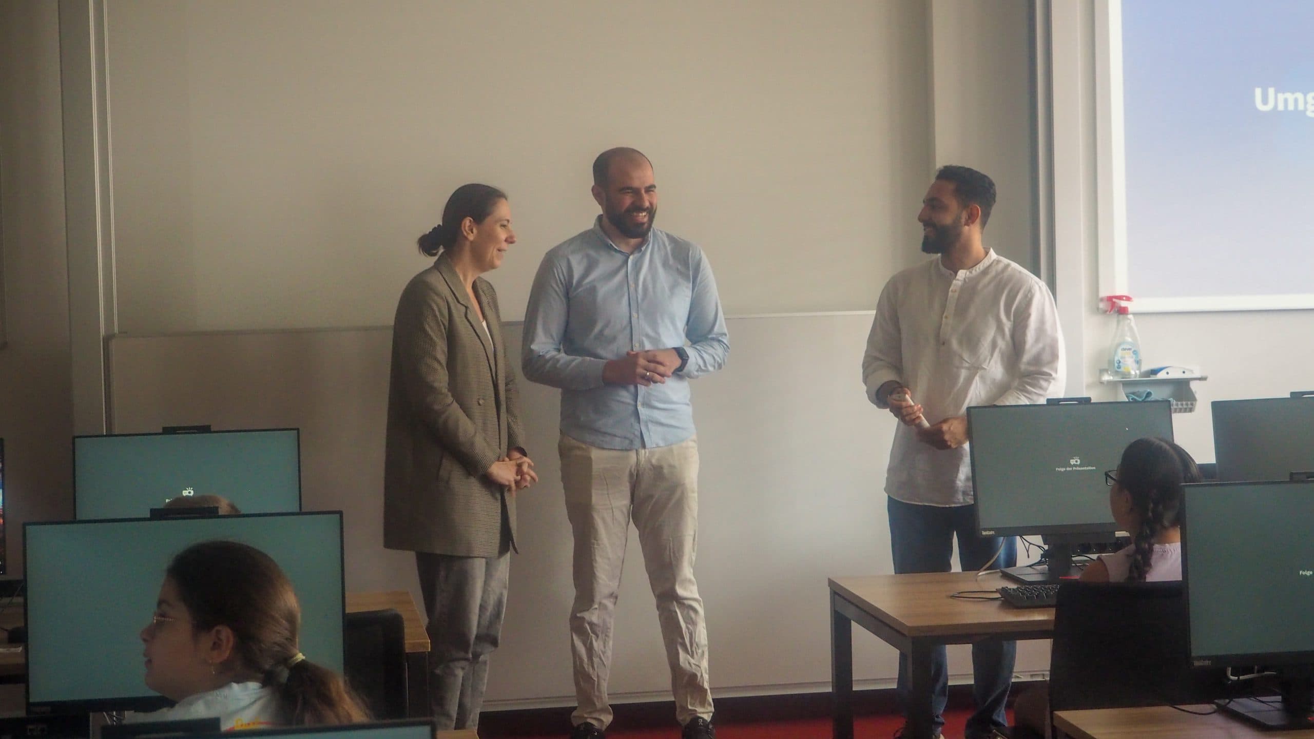 Drei Erwachsene stehen vor einem Computerlabor, lächeln und sprechen miteinander, während die Schüler an den Tischen sitzen und Computer benutzen. Im Hintergrund ist eine Projektionsfläche zu sehen.