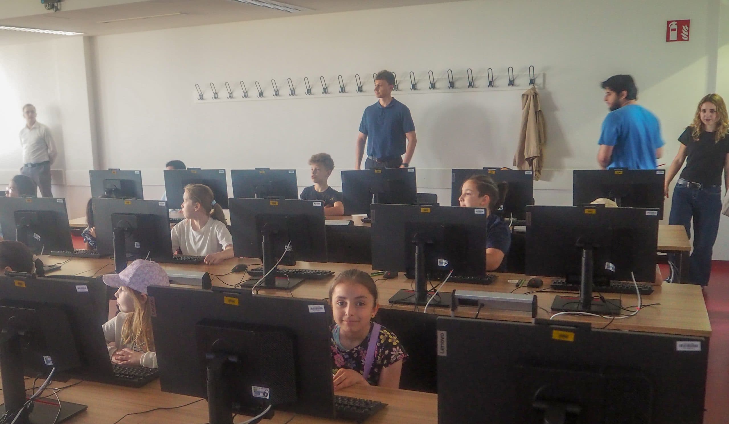 In einem Klassenzimmer sitzen Kinder an Computertischen und benutzen jeweils einen Computer. Mehrere Erwachsene stehen im hinteren und seitlichen Teil des Raumes und schauen zu. Der Raum ist gut beleuchtet, hat helle Wände und Kleiderhaken an der Wand.