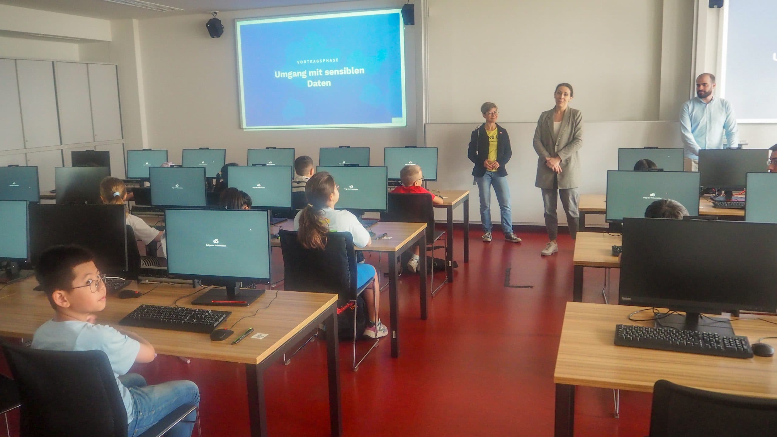 Ein Klassenzimmer mit jungen Schülern, die an Computern sitzen und auf einen Bildschirm mit der Aufschrift 'Umgang mit sensiblen Daten' schauen. Vorne stehen drei Erwachsene, die sich darauf vorbereiten, über den Umgang mit sensiblen Daten zu unterrichten.