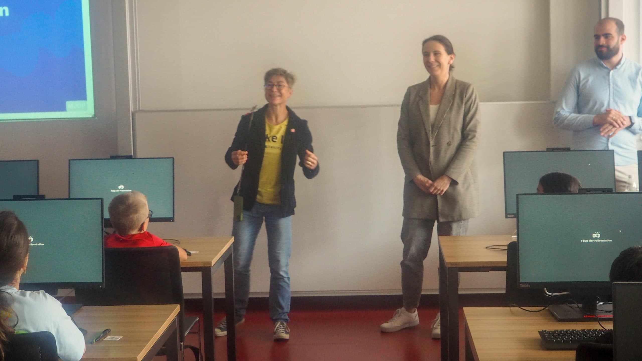 Drei Erwachsene stehen vorne in einem Klassenzimmer und halten eine Präsentation vor Schülern, die an Tischen mit Computermonitoren sitzen. Der Raum ist mit einem Whiteboard und einer an die Wand projizierten Präsentation ausgestattet.