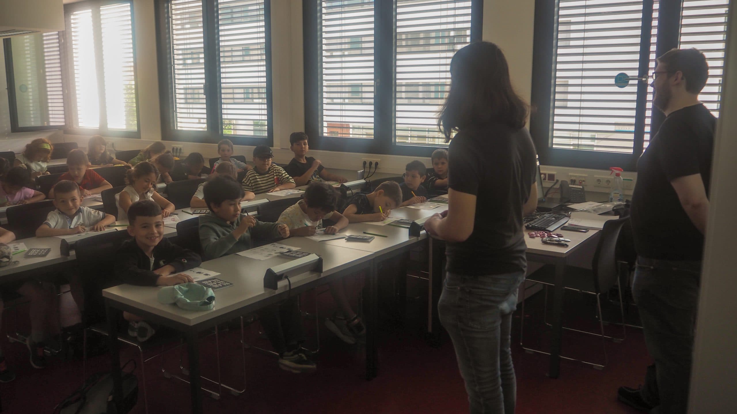 Ein Klassenzimmer mit Kindern, die an Tischen sitzen, einige schreiben oder zeichnen, während zwei Erwachsene im vorderen Teil des Raumes neben einem Tisch mit Papieren und einem Computer stehen. Das Sonnenlicht fällt durch große Fenster mit Jalousien.