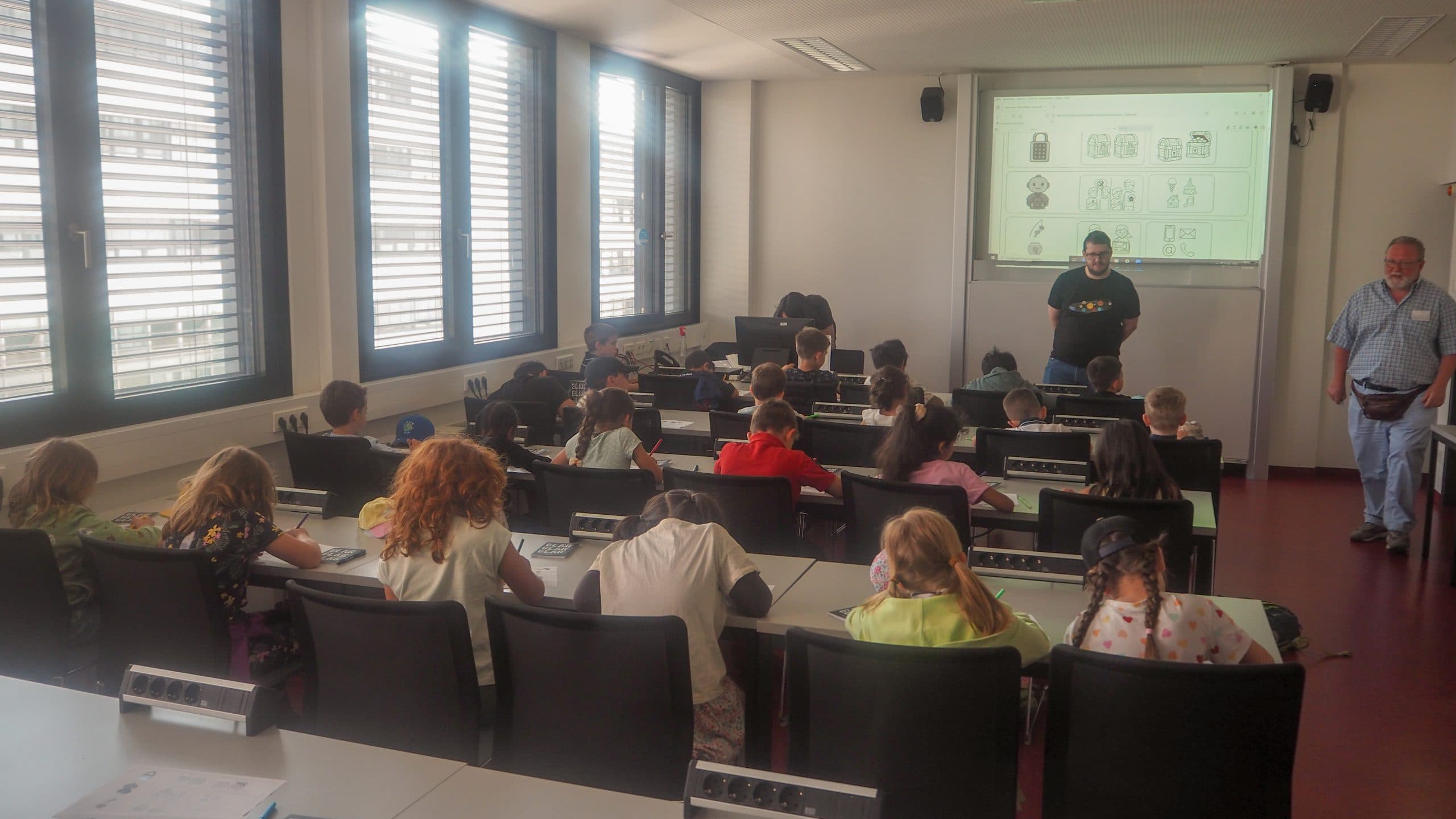 Ein Klassenzimmer mit jungen Schülern, die an den Tischen sitzen und zwei Erwachsenen zuhören, die vorne sitzen. Ein Erwachsener steht neben einer projizierten Präsentation, während der andere an der Seite steht. Das Sonnenlicht fällt durch große Fenster auf der linken Seite ein.