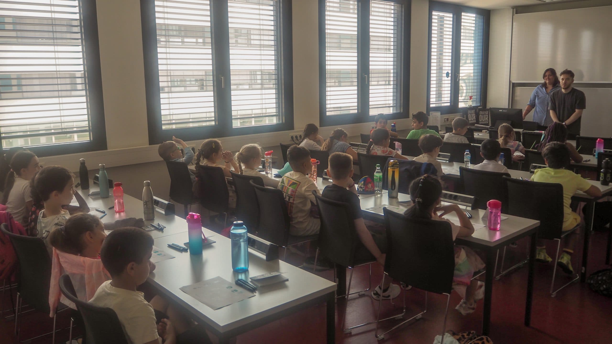 Ein Klassenzimmer mit jungen Schülern, die an Tischen sitzen, jeder mit einer Wasserflasche, gegenüber zwei Erwachsenen an der Stirnseite. Große Fenster mit Jalousien lassen natürliches Licht herein, und das Klassenzimmer hat eine helle, aufmerksame Atmosphäre.