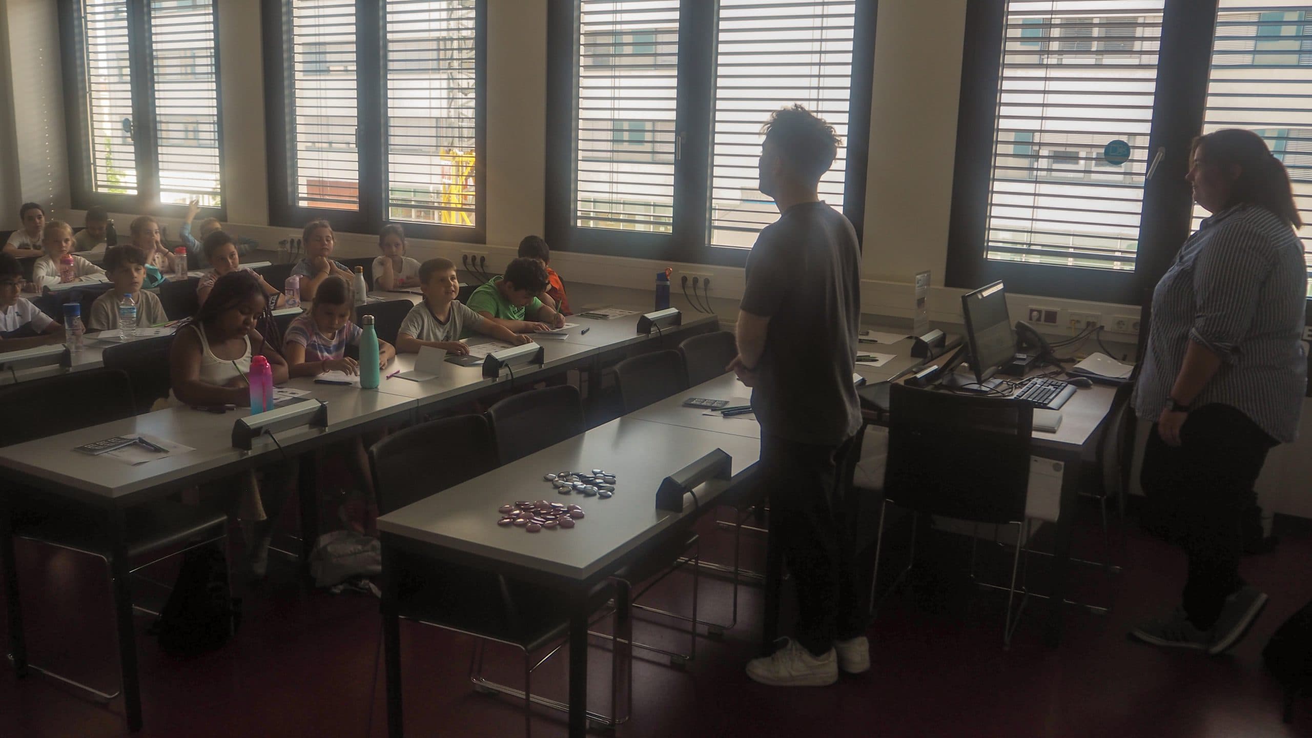 Eine Gruppe von Kindern sitzt in einem Klassenzimmer an den Tischen und blickt auf zwei Erwachsene, die an der Stirnseite neben einem Computer stehen. Sonnenlicht strömt durch große Fenster im Hintergrund.