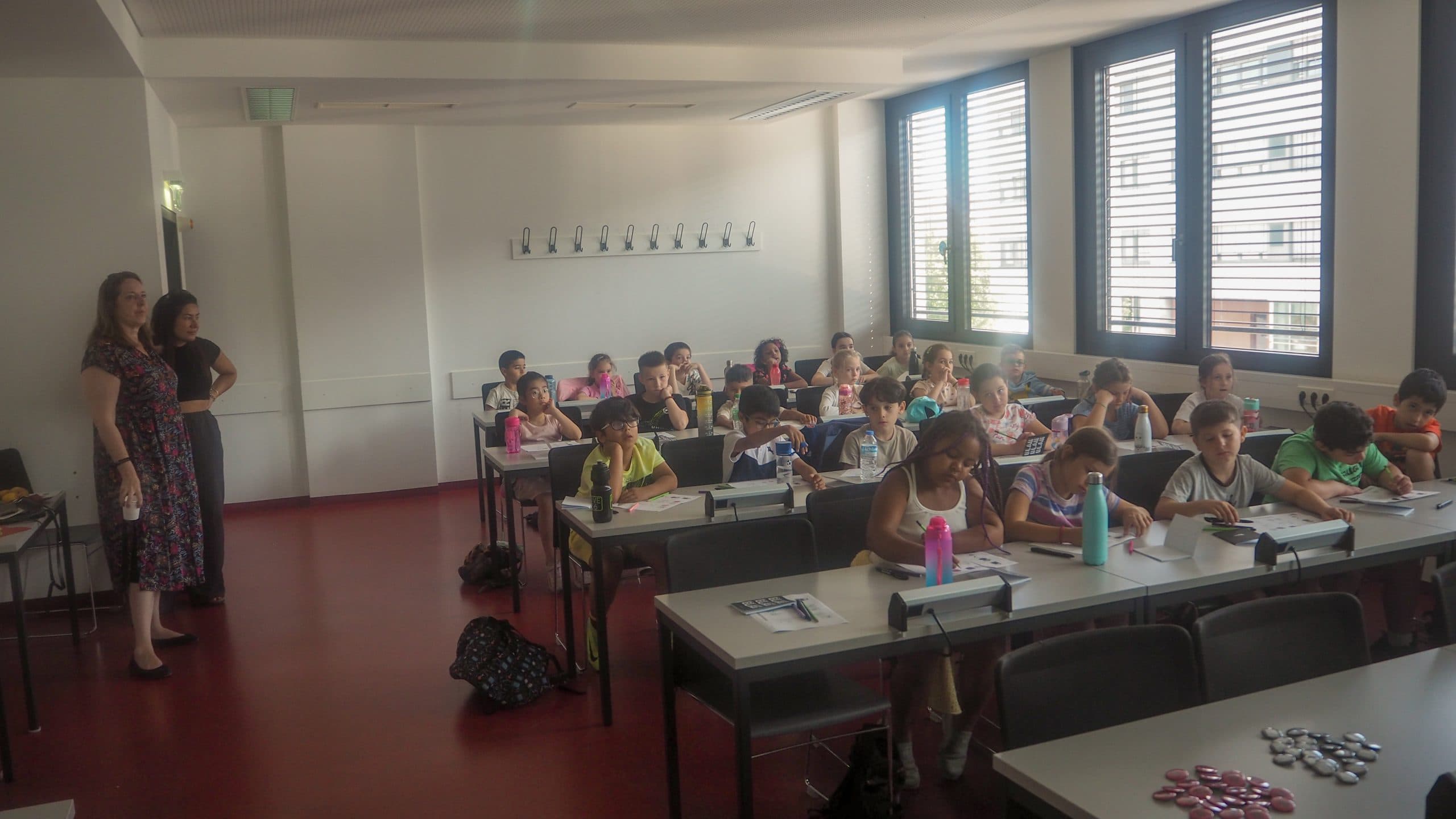 Ein Klassenzimmer mit zwei Lehrern, die vorne stehen, und mehreren Kindern, die an Tischen sitzen und teilweise schreiben oder zeichnen. Der Raum ist hell, hat große Fenster und einen roten Fußboden.