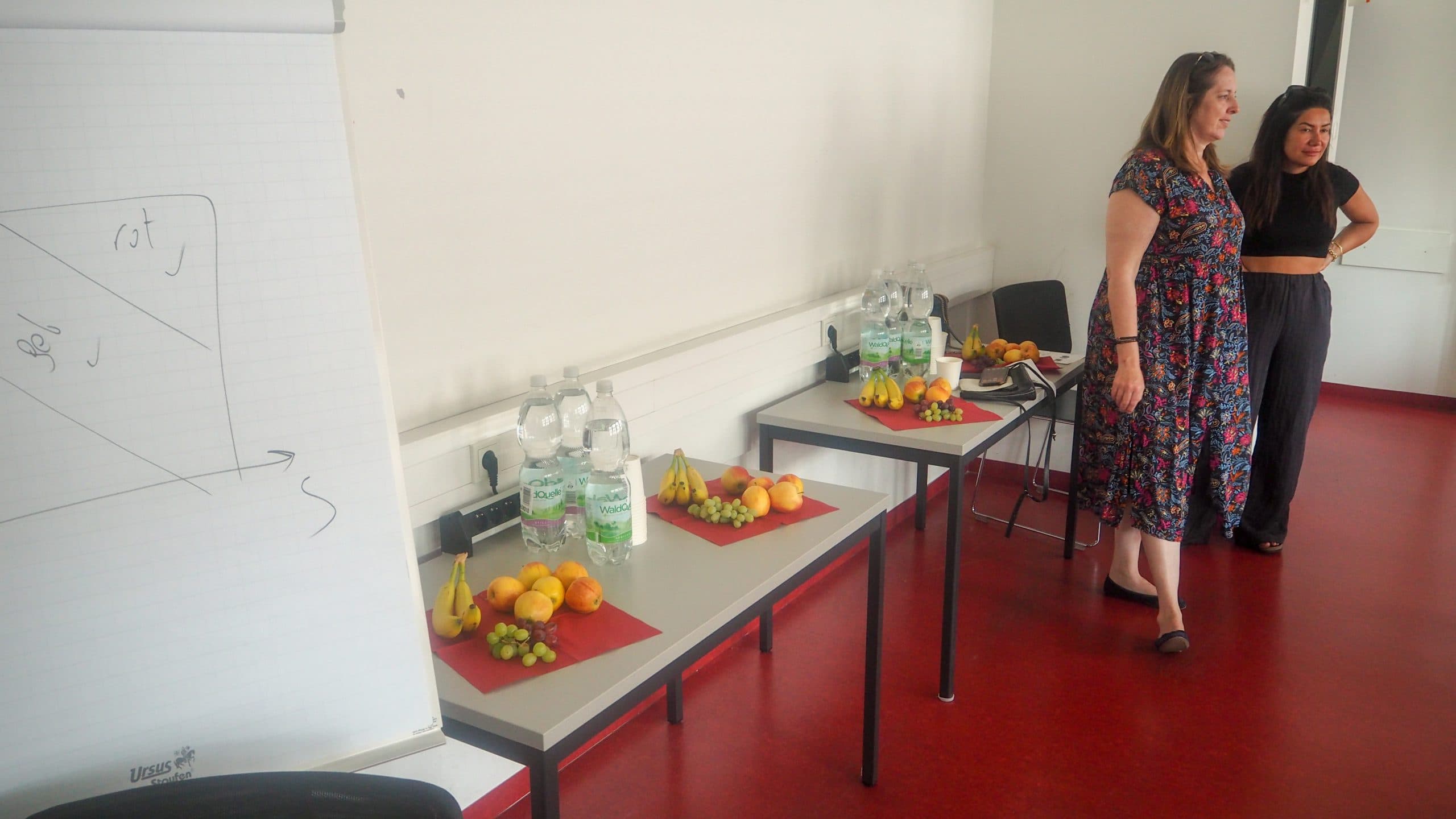 Zwei Frauen stehen in einem Klassenzimmer mit rotem Bodenbelag. Auf zwei Tischen stehen Wasserflaschen, Gläser und Obstteller. Auf der linken Seite des Bildes ist ein Flipchart mit einer einfachen Grafik zu sehen.