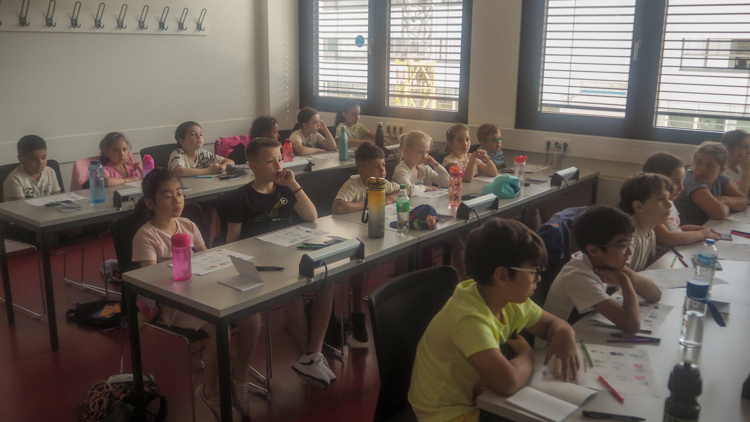 Ein Klassenzimmer mit aufmerksamen Kindern sitzt an den Tischen und hört einer Unterrichtsstunde zu. Die Schüler haben Hefte, Wasserflaschen und verschiedene Materialien auf ihren Tischen. Das Sonnenlicht fällt durch die großen Fenster hinter ihnen ein.