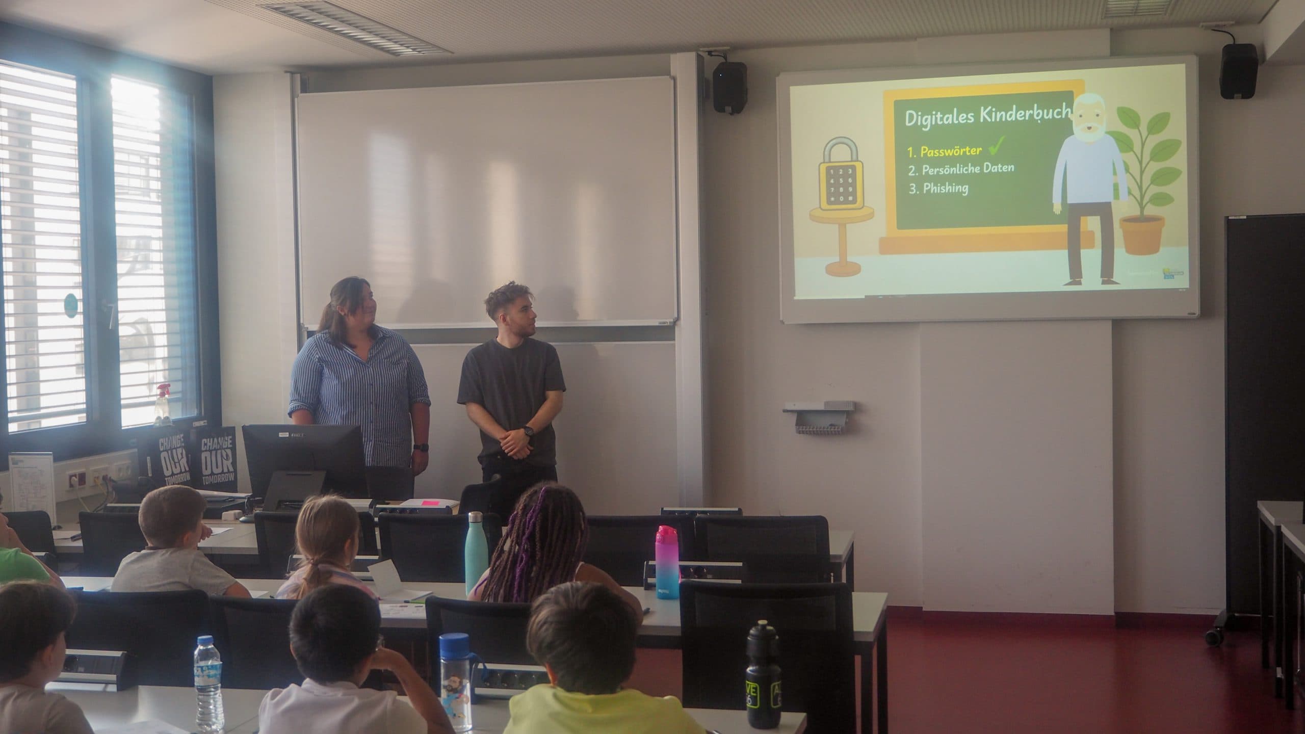 Zwei Erwachsene stehen vor einem Klassenzimmer mit Kindern und präsentieren eine Folie mit dem Titel Digitales Kinderbuch mit drei Aufzählungspunkten. Im Klassenzimmer sitzen die Schüler an den Tischen und schauen in Richtung der Vortragenden.