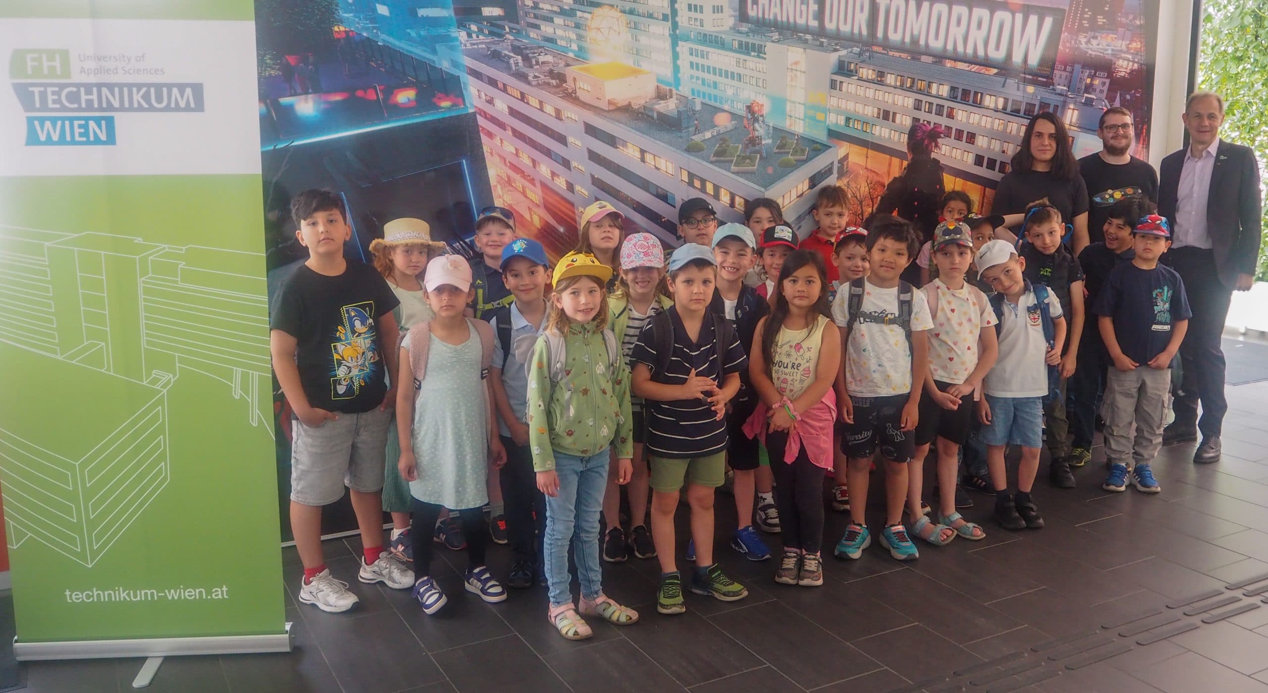 Eine Gruppe kleiner Kinder steht vor einem großen Plakat und einem grünen Werbebanner der FH Technikum Wien und posiert für ein Foto mit zwei Erwachsenen an beiden Enden. Die Kinder tragen Rucksäcke, Mützen und legere Sommerkleidung.