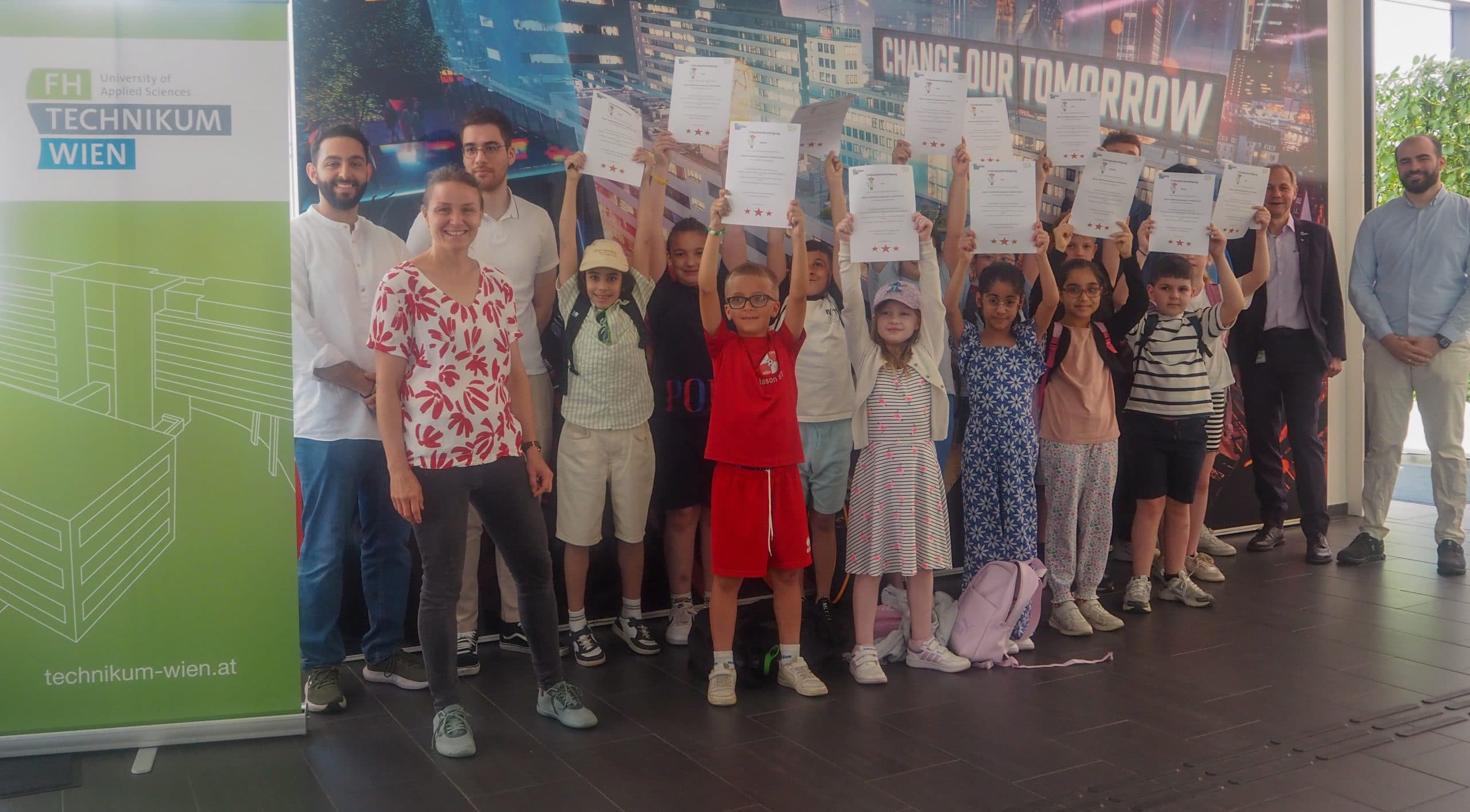 Eine Gruppe von Kindern hält stolz ihre Zertifikate hoch, während sie mit mehreren Erwachsenen vor einem Banner und einem grünen Technikum Wien-Schild stehen. Alle lächeln, und Rucksäcke liegen auf dem Boden.