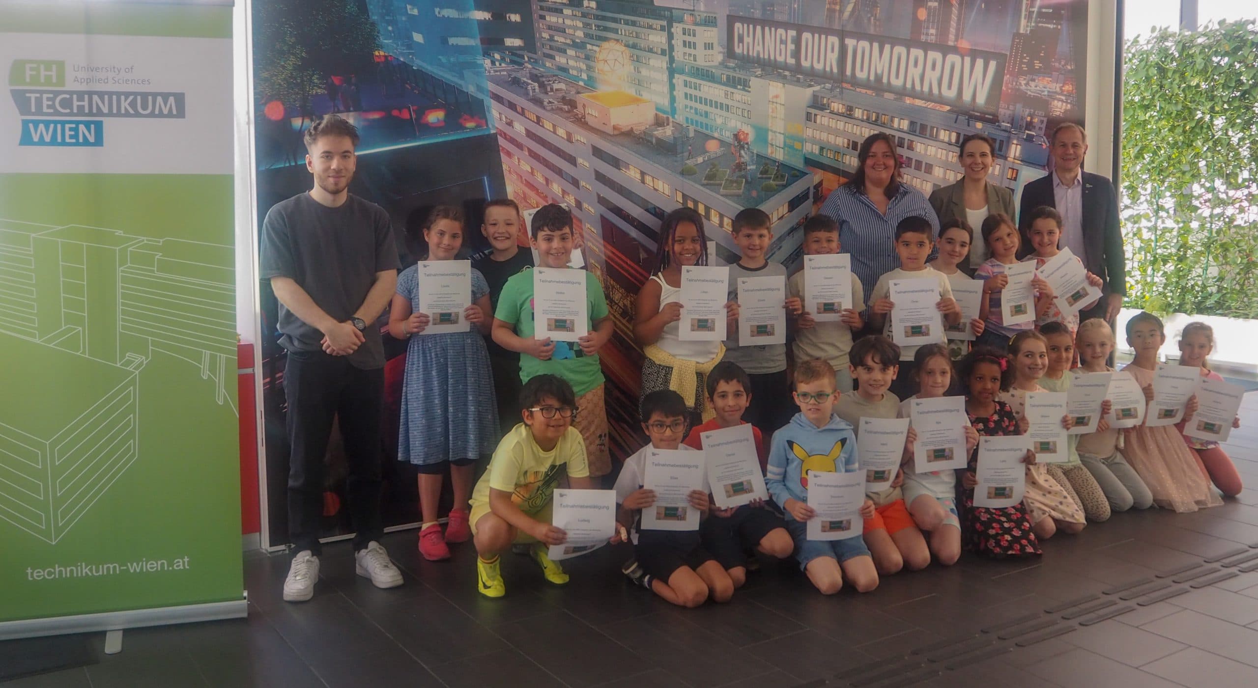 Eine Gruppe von Kindern und mehrere Erwachsene posieren in einem Innenraum mit Urkunden vor einem Transparent mit der Aufschrift Change Our Tomorrow und einem Schild der FH Technikum Wien. Alle lächeln in die Kamera.