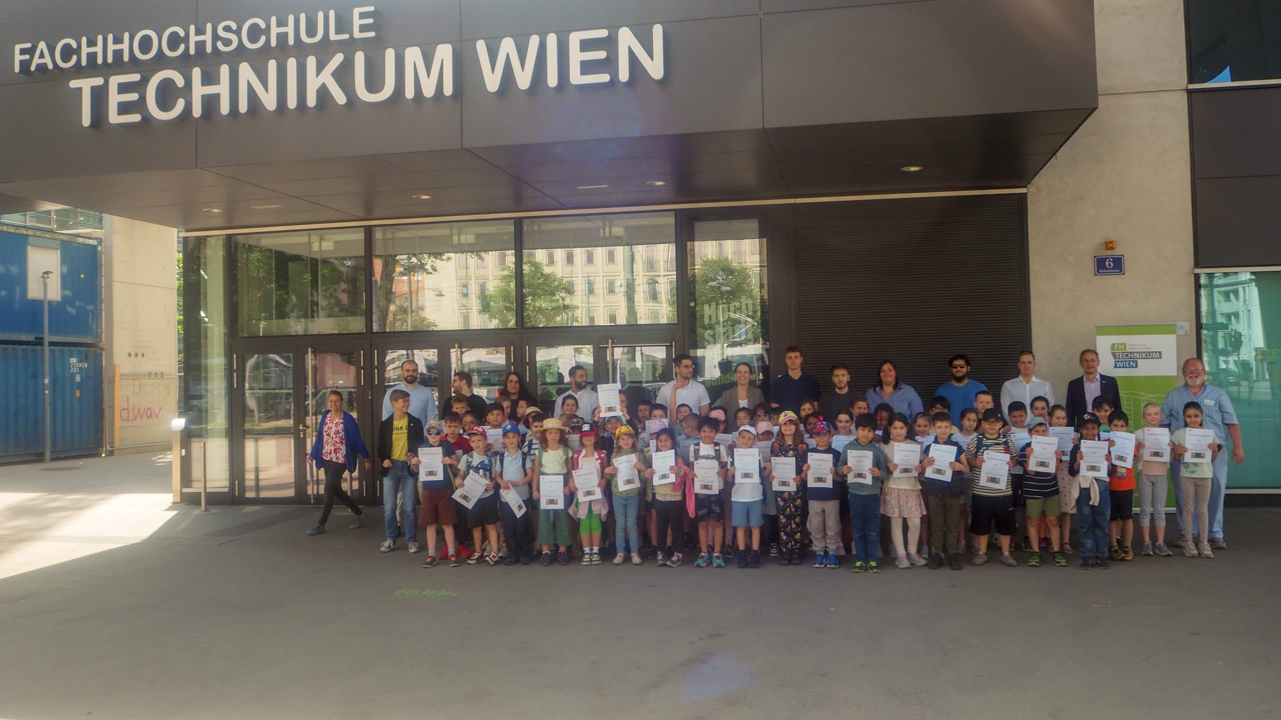 Eine große Gruppe von Kindern mit Urkunden posiert mit mehreren Erwachsenen vor dem Eingang der Fachhochschule Technikum Wien, einem Universitätsgebäude in Wien.