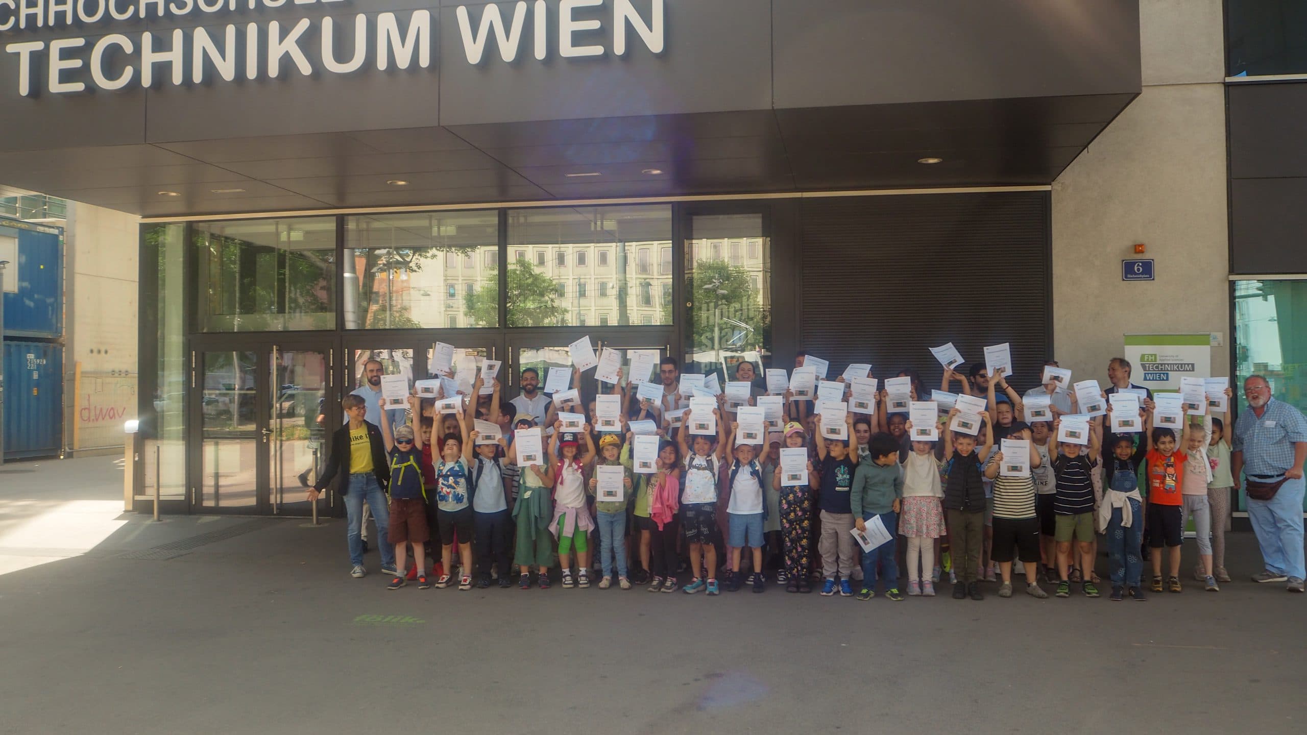 Eine große Gruppe von Kindern und Erwachsenen steht vor einem Gebäude und hält lächelnd Urkunden hoch. Auf dem Gebäudeschild steht TECHNIKUM WIEN. Es ist ein sonniger Tag.
