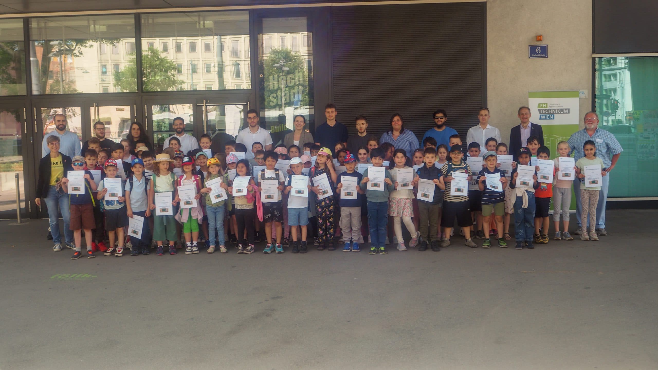 Eine große Gruppe von Kindern mit Urkunden posiert mit mehreren Erwachsenen vor einem modernen Gebäude mit Glasfenstern. Einige Kinder tragen Hüte und Sonnenbrillen, und alle lächeln.